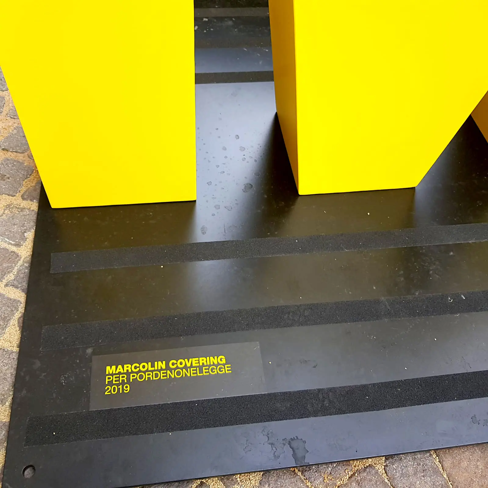 Strutture rettangolari gialle su base nera con il testo "MARCOLIN COVERING PER PORDENONELEGGE 2019" su una targa nell'angolo inferiore. La superficie è su un sentiero di ciottoli.