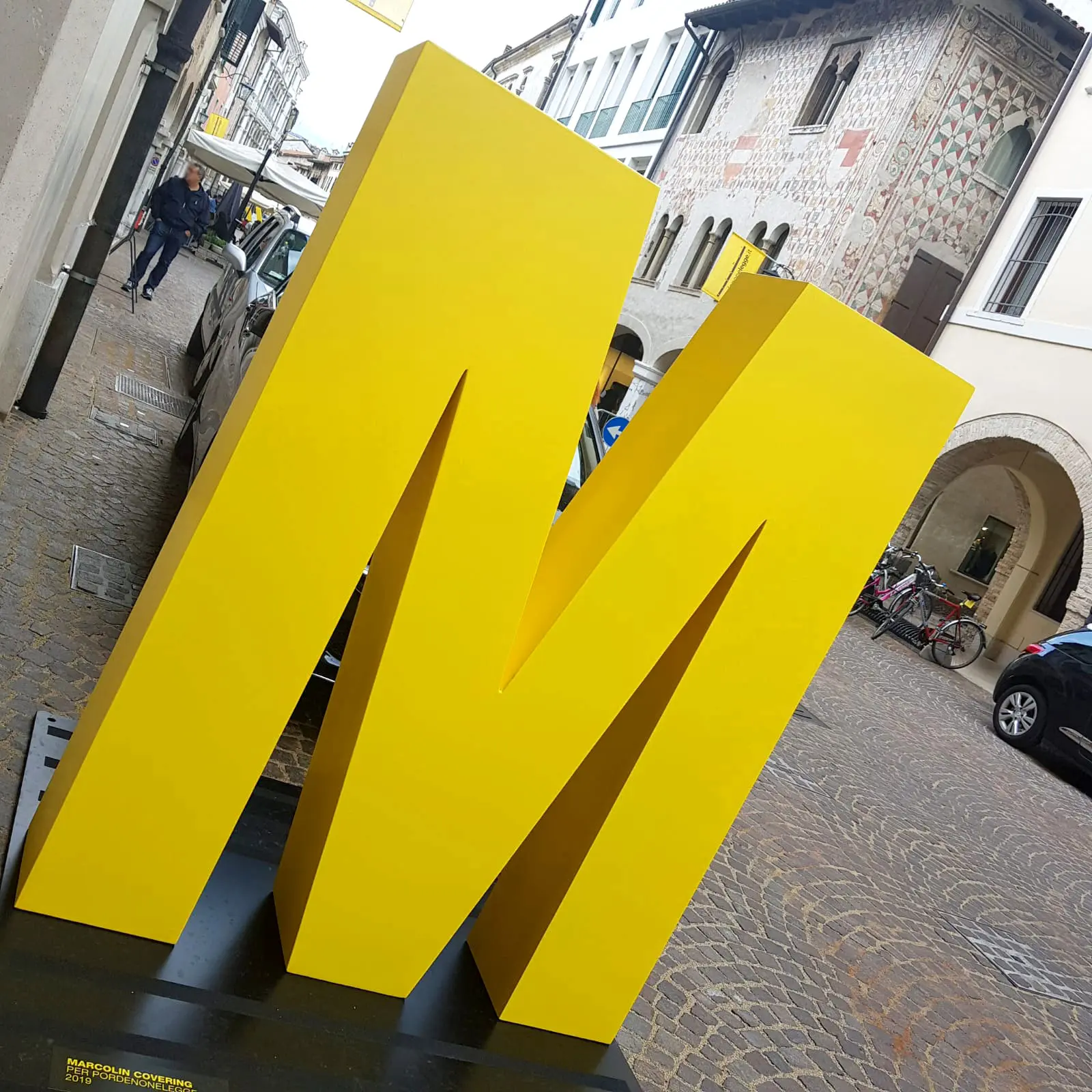 Una grande scultura gialla brillante raffigurante la lettera "M" si trova su una strada acciottolata. Sullo sfondo, ci sono edifici con archi, persone che camminano e biciclette parcheggiate.
