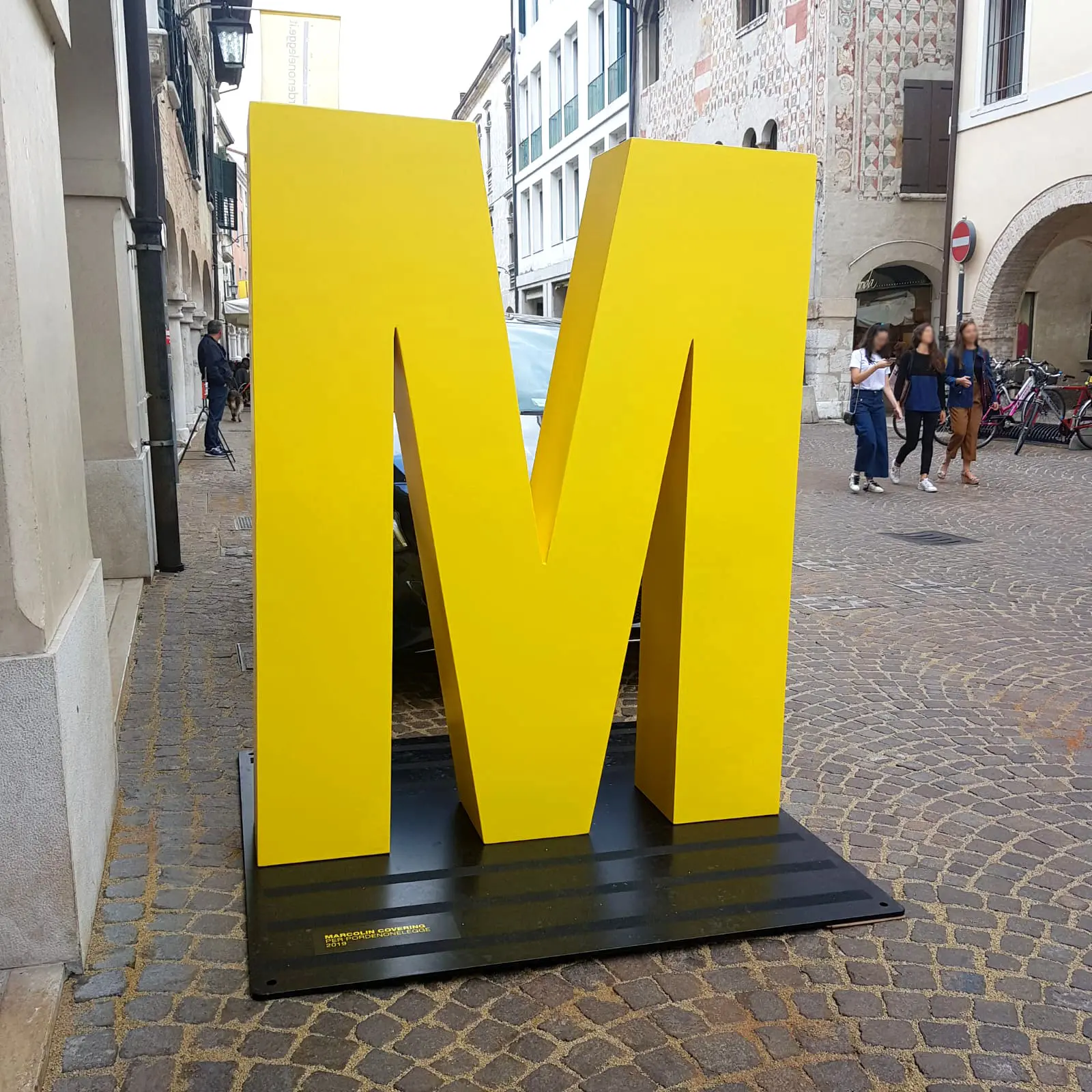 Una grande scultura gialla a forma di lettera "M" si erge su una strada acciottolata. Sullo sfondo, le persone camminano e le biciclette sono parcheggiate lì vicino. Gli edifici che costeggiano la strada hanno uno stile architettonico europeo.