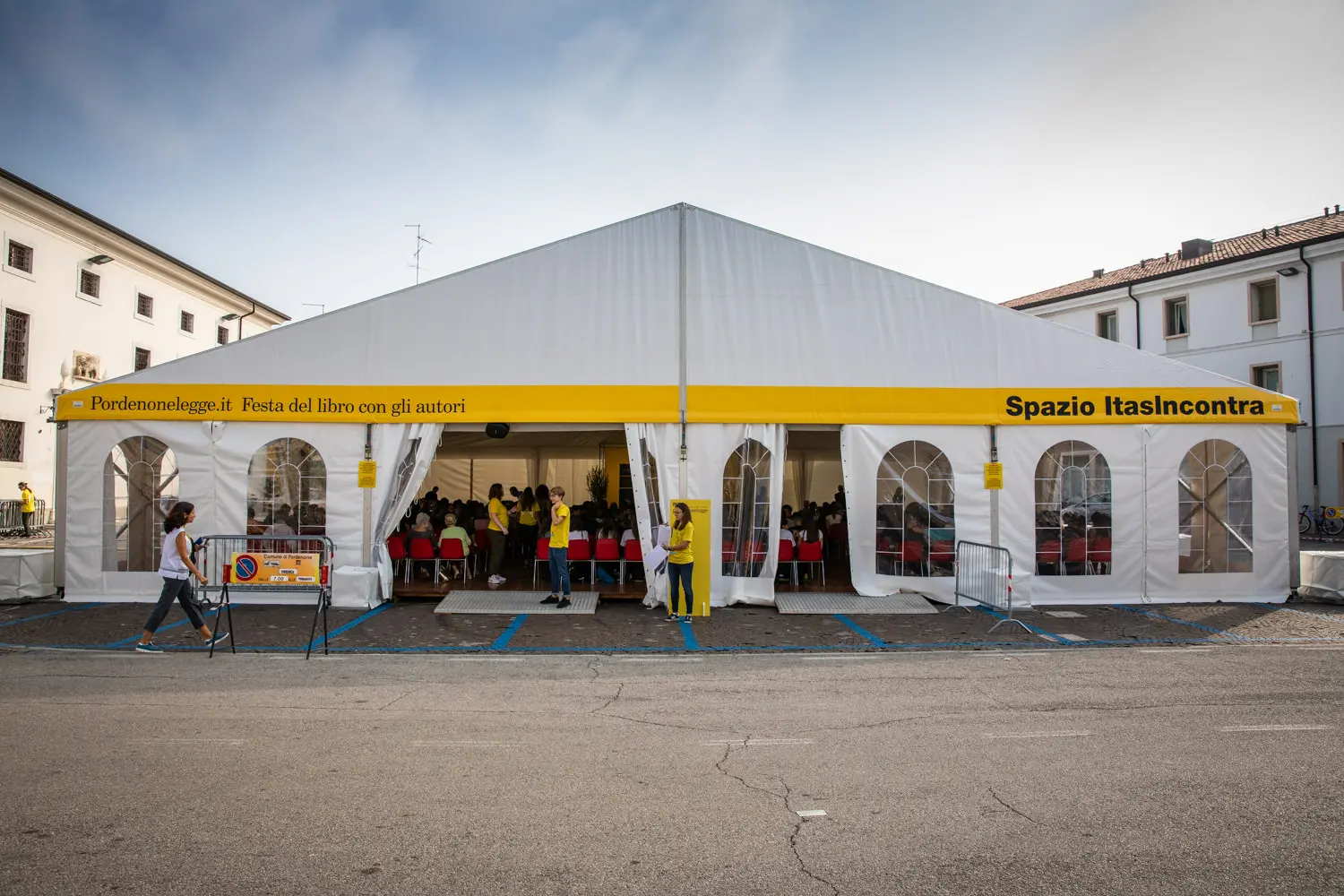 Una grande tenda bianca con la scritta "Spazio ItasIncontra" con persone visibili all'interno, parte del festival del libro Pordenonelegge. La tenda ha accenti e striscioni gialli. La scena è ambientata in un'area pavimentata con edifici bianchi sullo sfondo.
