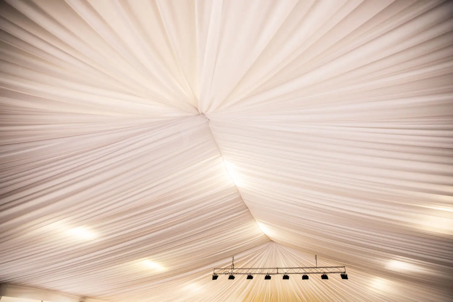 Un soffitto con tessuto bianco drappeggiato, che crea un aspetto simile a una tenda. Quattro faretti su una struttura metallica sono appesi centralmente, proiettando una luce calda sul tessuto, creando un'atmosfera morbida ed elegante.