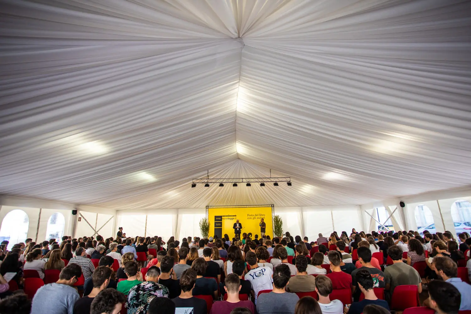 Un folto gruppo di persone sedute all'interno di una tenda bianca di fronte a un palco giallo con un oratore a un leggio. Il soffitto è alto con tessuto drappeggiato e ci sono grandi finestre che lasciano entrare la luce naturale.