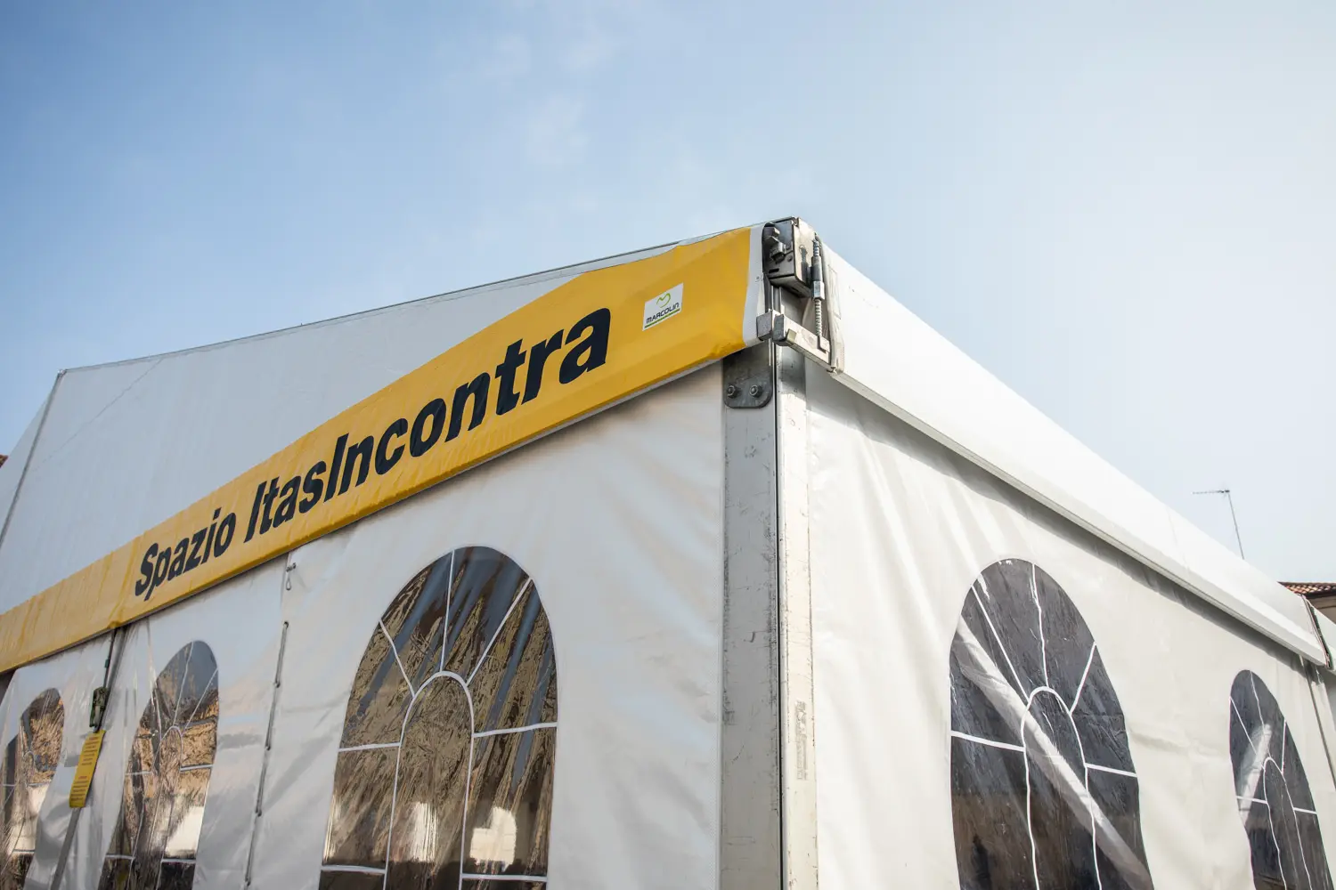 Una grande tenda bianca con finestre ad arco e un cartello giallo all'angolo con la scritta "Spazio ItasIncontra". Il cielo è limpido e azzurro sullo sfondo.