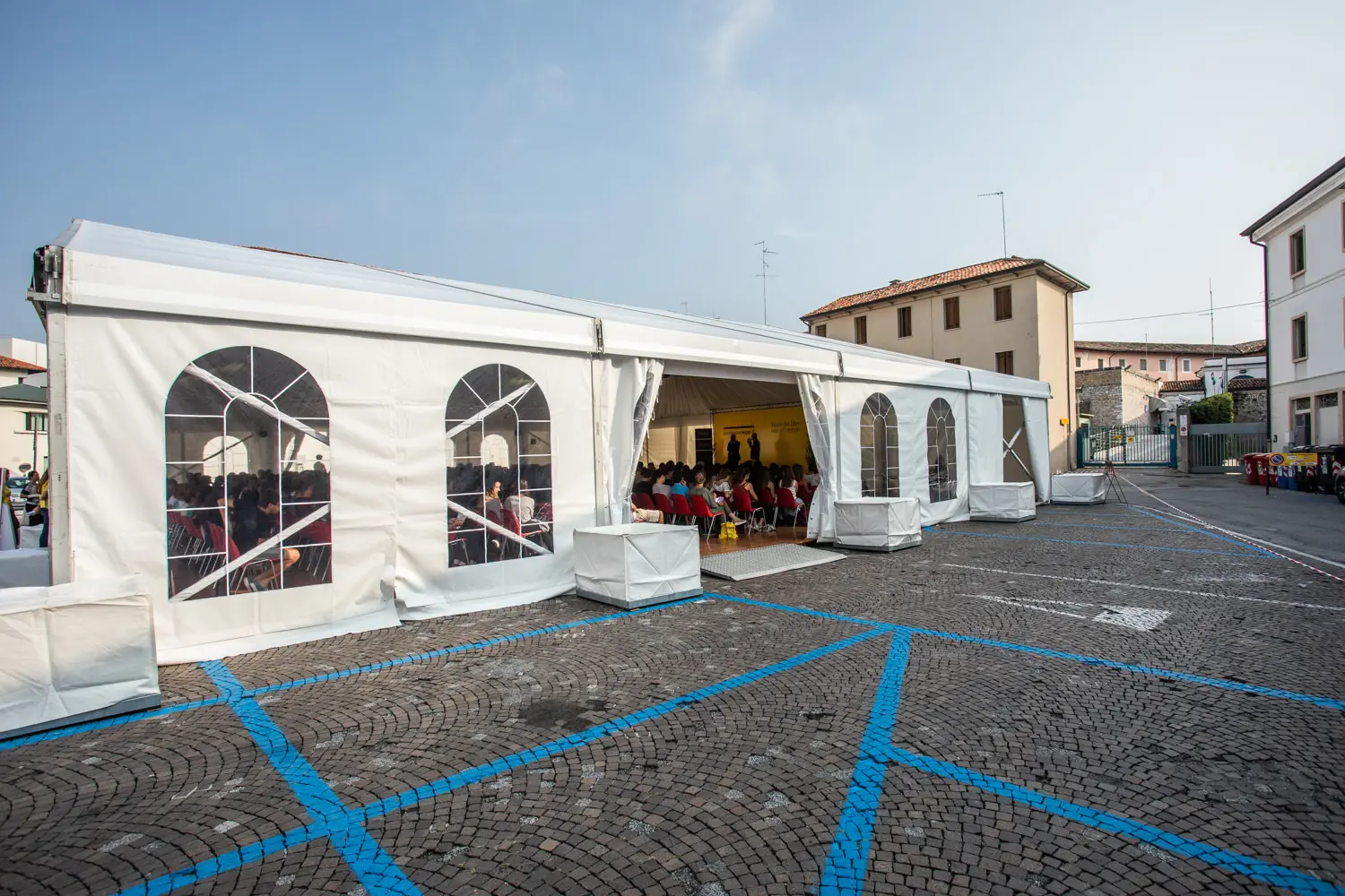 Una grande tenda bianca con finestre ad arco è installata su un'area acciottolata. All'interno, le persone sono sedute di fronte a un palco. Il terreno esterno è segnato da linee blu, che probabilmente indicano i parcheggi. Gli edifici sono visibili sullo sfondo sotto un cielo limpido.