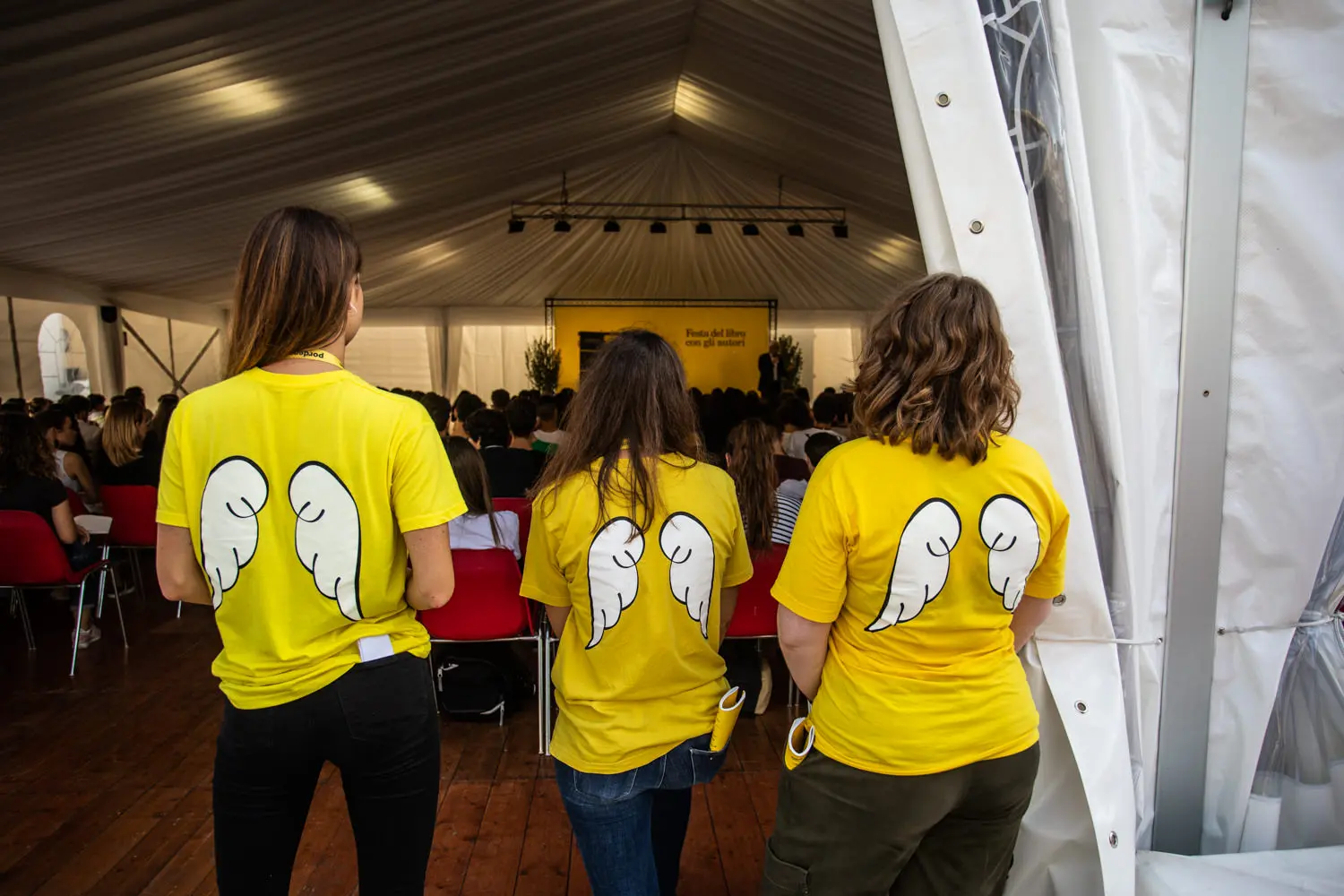 Tre persone che indossano magliette gialle con disegni di ali bianche sono in piedi di fronte a un raduno affollato all'interno di una grande tenda. Uno striscione giallo è appeso di fronte e l'area è ben illuminata, creando un'atmosfera vibrante.