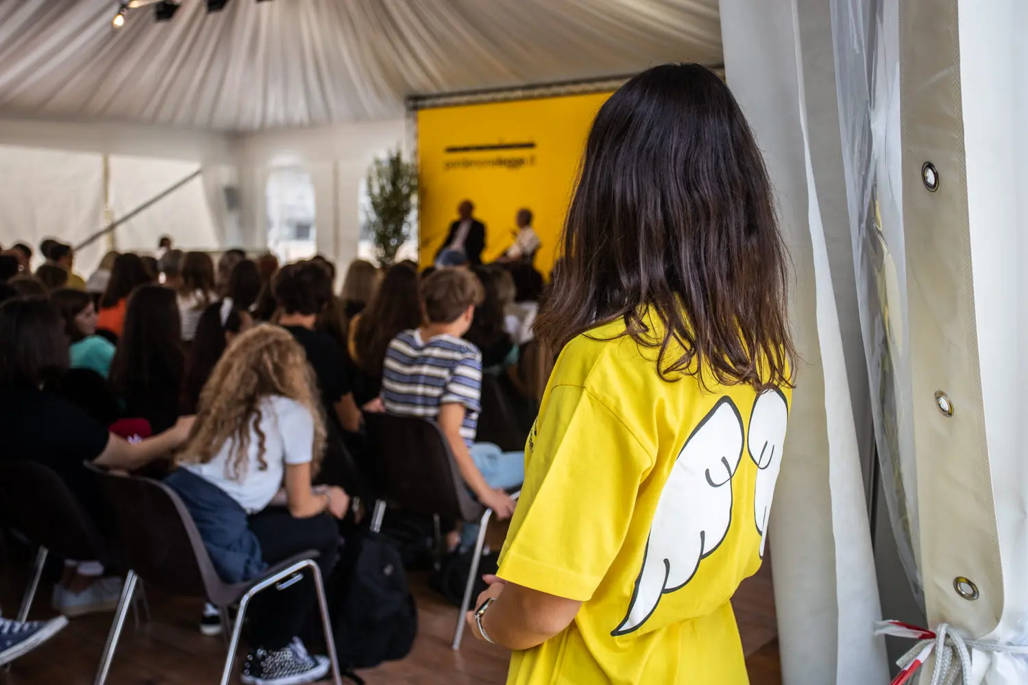 Una persona che indossa una maglietta gialla brillante con ali d'angelo bianche sulla schiena è in piedi vicino all'ingresso di una tenda. All'interno, le persone sono sedute, e guardano attentamente una presentazione sul palco. Lo sfondo è giallo con la parola "ancestramage" visibile.