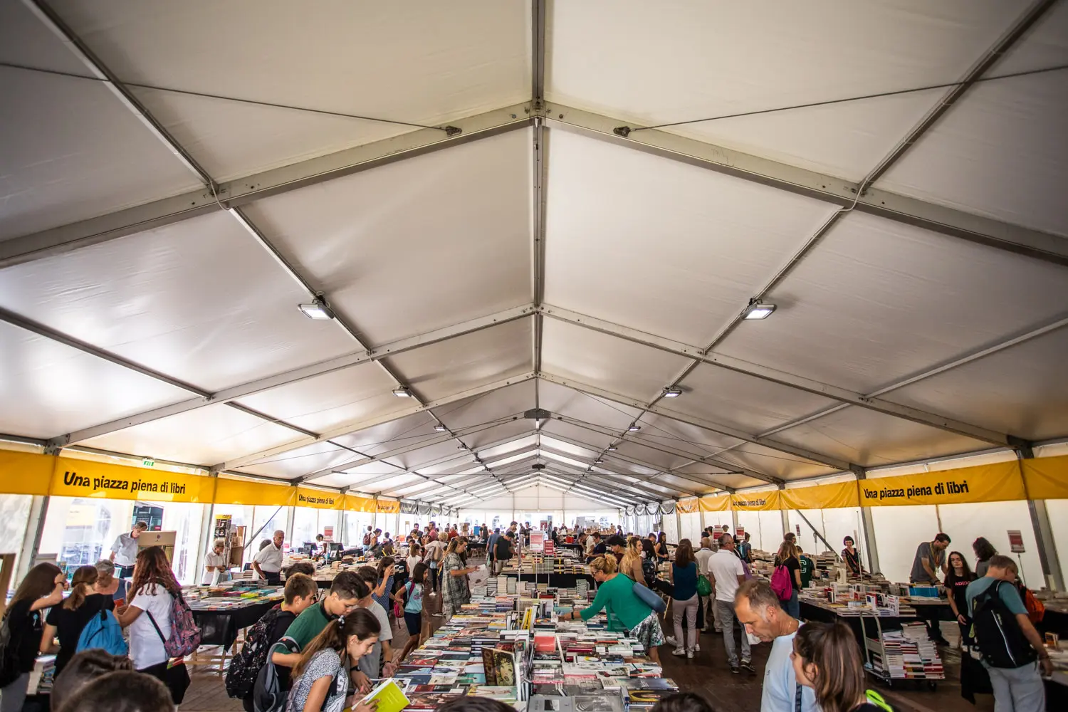 Un tendone bianco, grande e affollato, ospita una vivace fiera del libro. Le persone curiosano tra numerosi tavoli pieni di libri. All'interno sono appesi striscioni gialli con testo e l'atmosfera è vivace e coinvolgente.