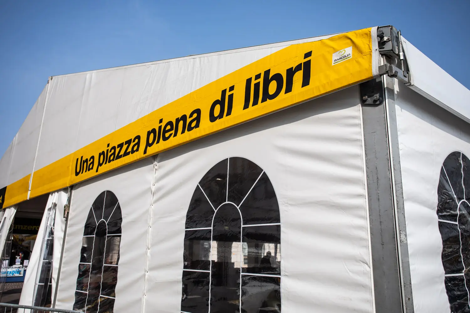 Tenda bianca con finestre ad arco e uno striscione giallo con la scritta in italiano "Una piazza piena di libri" sul lato. Cielo azzurro sullo sfondo.