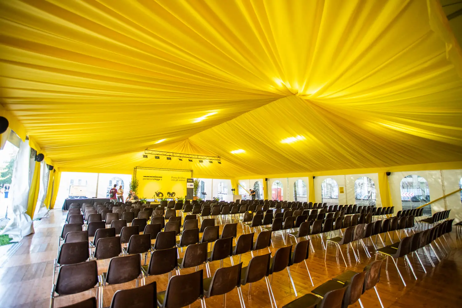 Un grande tendone per eventi è pieno di file di sedie marroni vuote di fronte a un palco. Il soffitto è drappeggiato con un tessuto giallo brillante e si possono vedere alcune persone che sistemano l'attrezzatura sul palco.