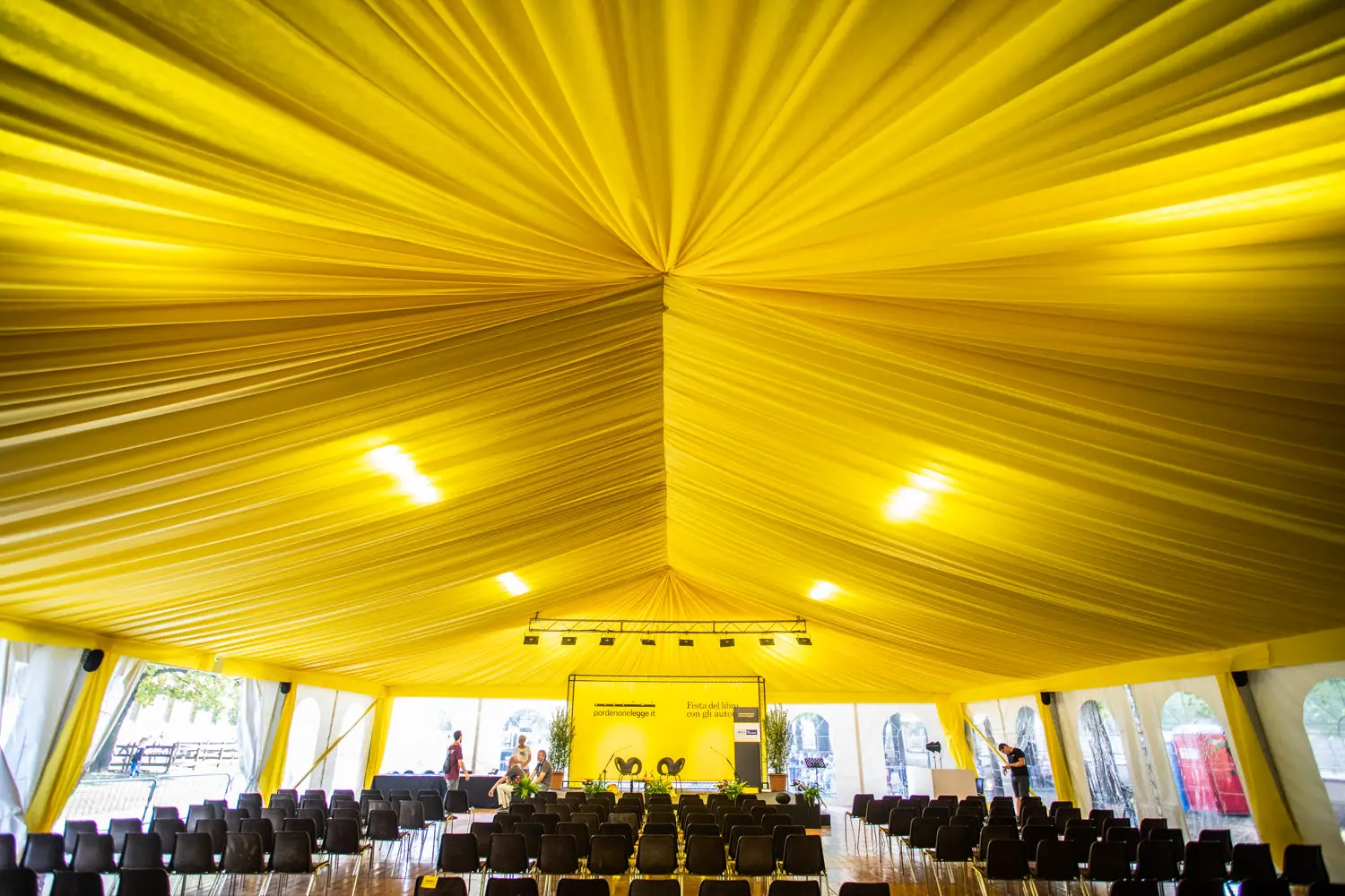 Una grande tenda con un soffitto giallo brillante e drappeggi in tessuto forma un baldacchino vibrante su file di sedie nere disposte di fronte a un palco. L'area del palco comprende uno sfondo giallo con luci e attrezzature allestite per un evento.