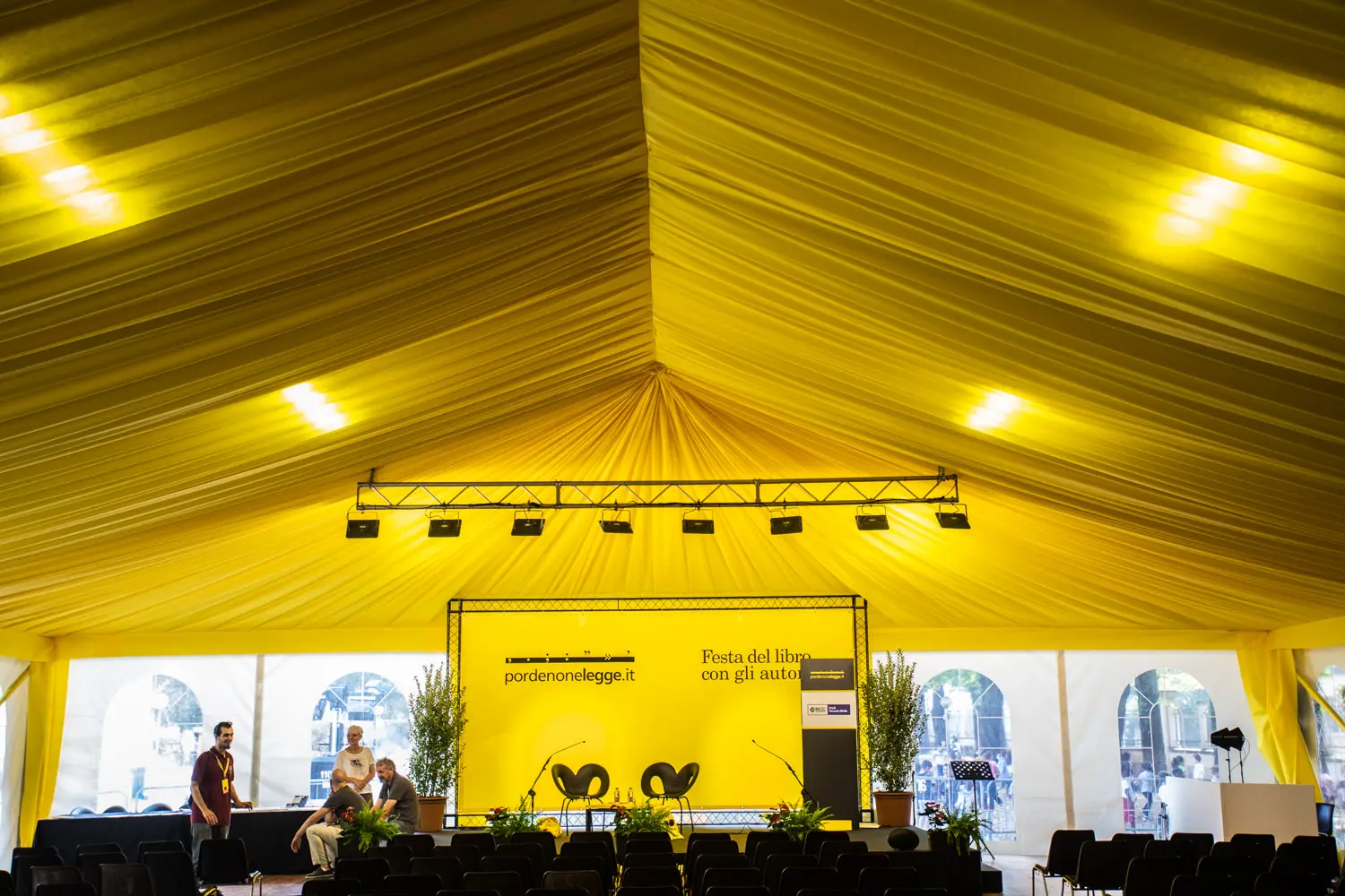 Un tendone da festival giallo brillante con un palco con due sedie gialle e uno sfondo con la scritta "Festa del libro con gli autori". Piante in vaso costeggiano il palco. Due persone stanno allestendo. File di sedie vuote sono rivolte verso il palco.