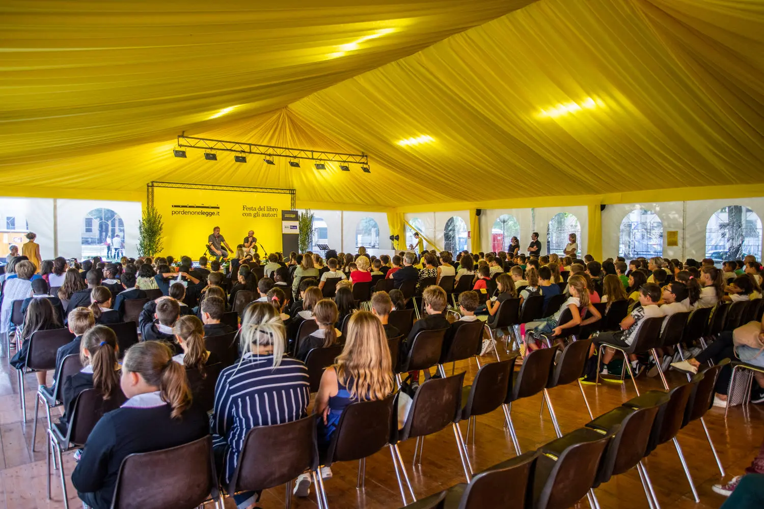 Un folto gruppo di persone sedute in fila all'interno di una tenda gialla, che guardano un gruppo di relatori su un palco. La tenda è luminosa e spaziosa e il pubblico comprende adulti e bambini. Luci sospese illuminano la scena.