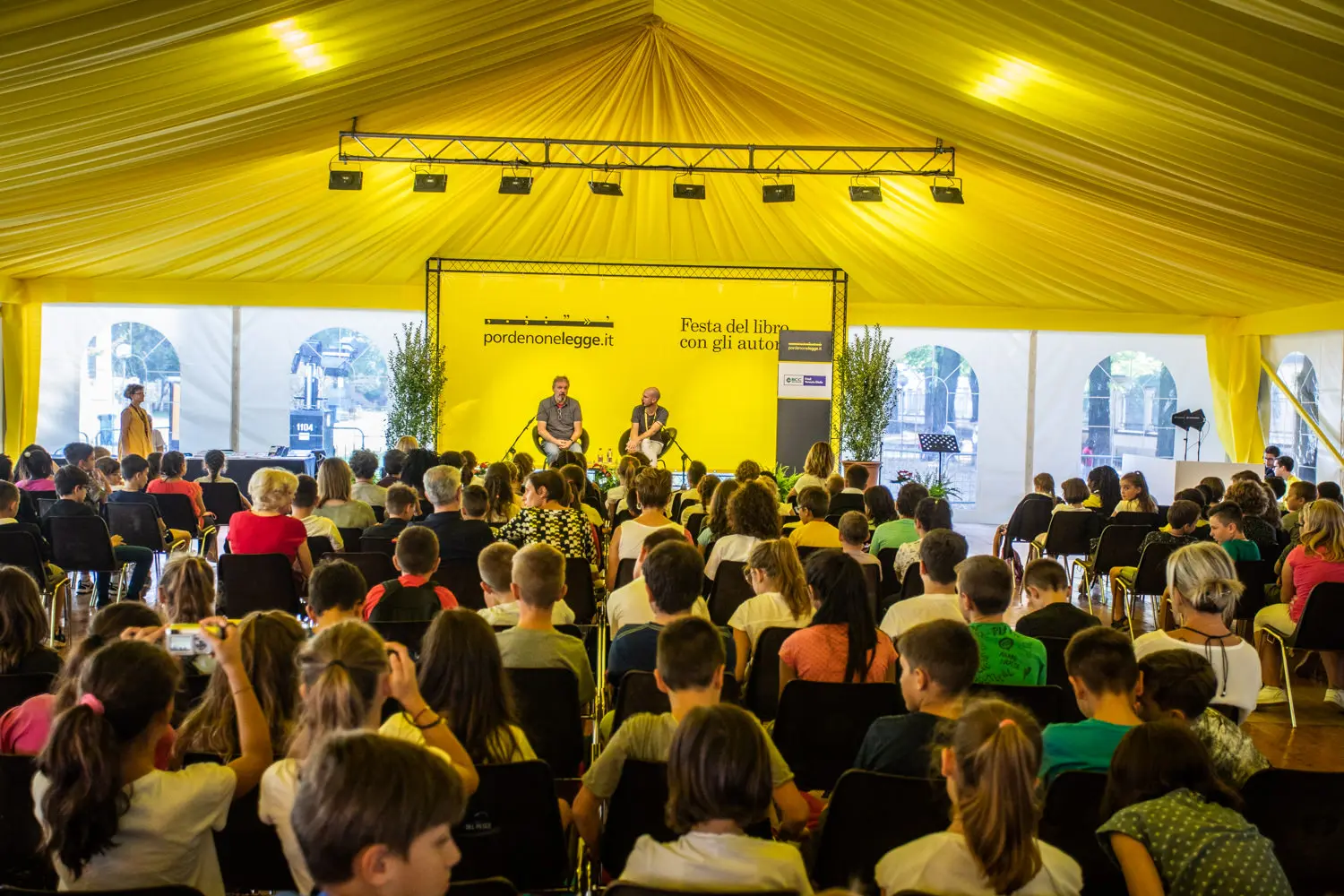Pubblico seduto in una grande tenda gialla che partecipa a un evento del festival del libro. Due relatori sono sul palco con uno sfondo giallo brillante che mostra i loghi dell'evento. L'atmosfera è vivace con molti bambini e adulti presenti.
