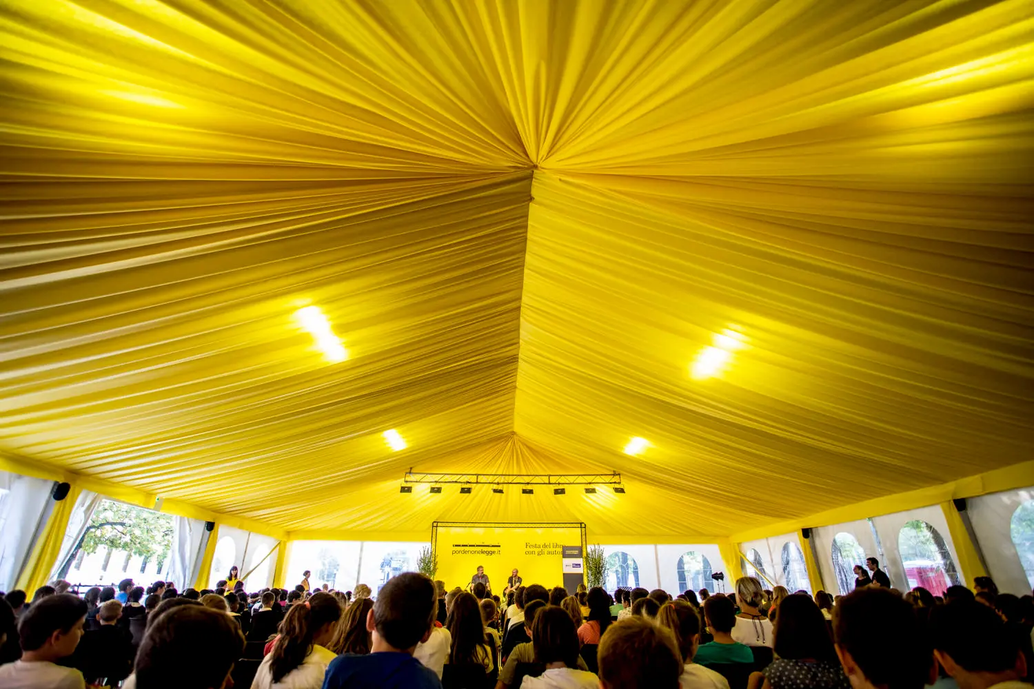 Una grande folla siede all'interno di una tenda gialla con soffitti drappeggiati, rivolta con attenzione verso un palco. Luci intense illuminano lo spazio e la luce naturale filtra attraverso i lati della tenda.