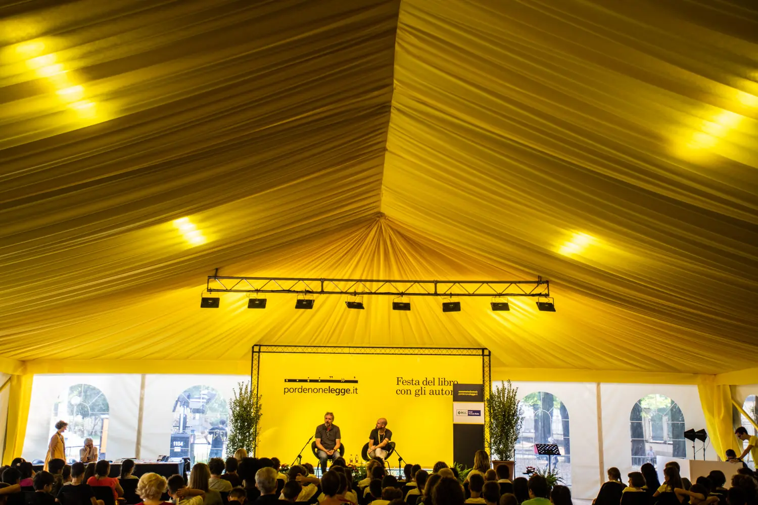 Una grande tenda gialla ospita un evento di festival del libro. Due relatori siedono sul palco con uno sfondo che recita "pordenonelegge.it" e "Festa del libro con gli autori". Il pubblico siede su sedie di fronte al palco e la luce naturale filtra attraverso i lati della tenda.