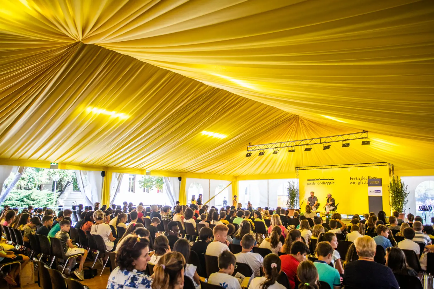 Un folto gruppo di persone sedute in fila all'interno di una tenda con soffitto e tende gialle. Due relatori sono sul palco e uno schermo dietro di loro mostra il nome dell'evento. Il pubblico è rivolto verso il palco e la tenda è illuminata intensamente.