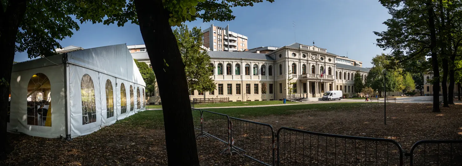 Vista panoramica di un edificio neoclassico con finestre ad arco, fiancheggiato da alberi. Nelle vicinanze si trova una tenda bianca con finestre ad arco. Sullo sfondo è visibile un edificio moderno e in primo piano un'area recintata.