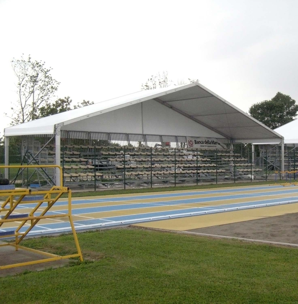 Una foto di un'area per eventi di atletica all'aperto con posti a sedere su tribune sotto una grande tenda bianca. Le tribune sono vuote e la pista ha corsie blu e gialle. Si possono vedere degli alberi sullo sfondo.