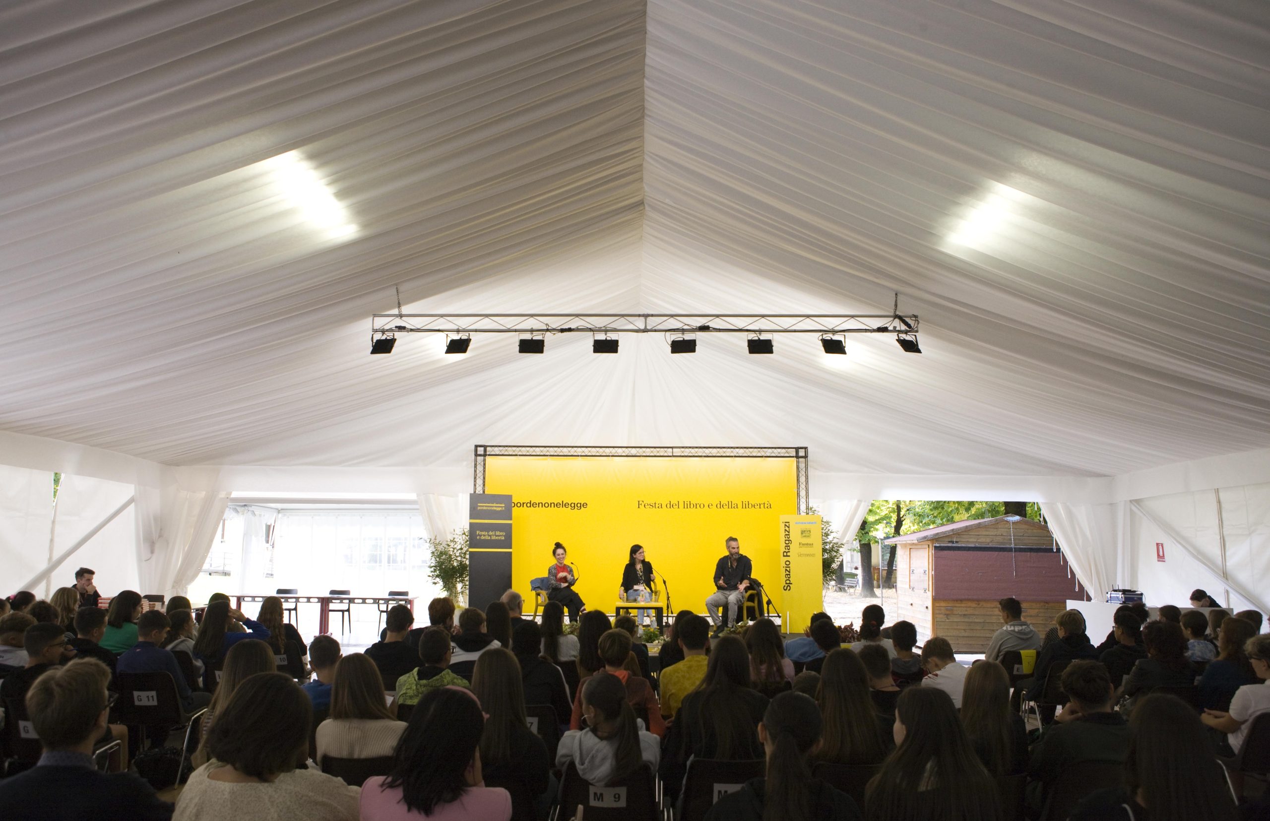 Un vasto pubblico siede in una tenda bianca di fronte a uno sfondo di palco giallo brillante. Tre persone sono sedute sul palco per una tavola rotonda. La luce naturale filtra attraverso le aperture della tenda e un cartello recita: "Liberi come il vento, liberi come i libri" in italiano.