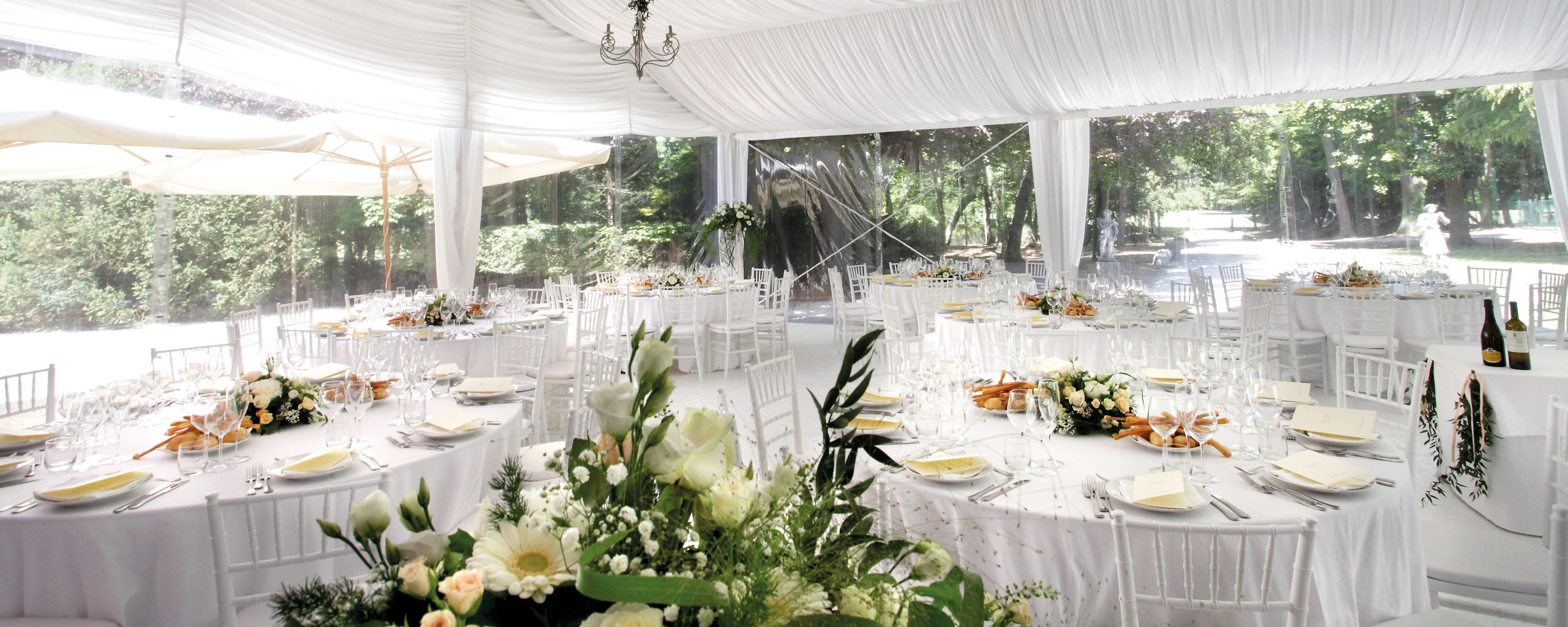 Un elegante ricevimento di nozze allestito sotto un baldacchino bianco. I tavoli rotondi sono adornati con tovaglie bianche, centrotavola floreali e segnaposto. Un lampadario pende dal soffitto e il verde è visibile attraverso i lati trasparenti della tenda.