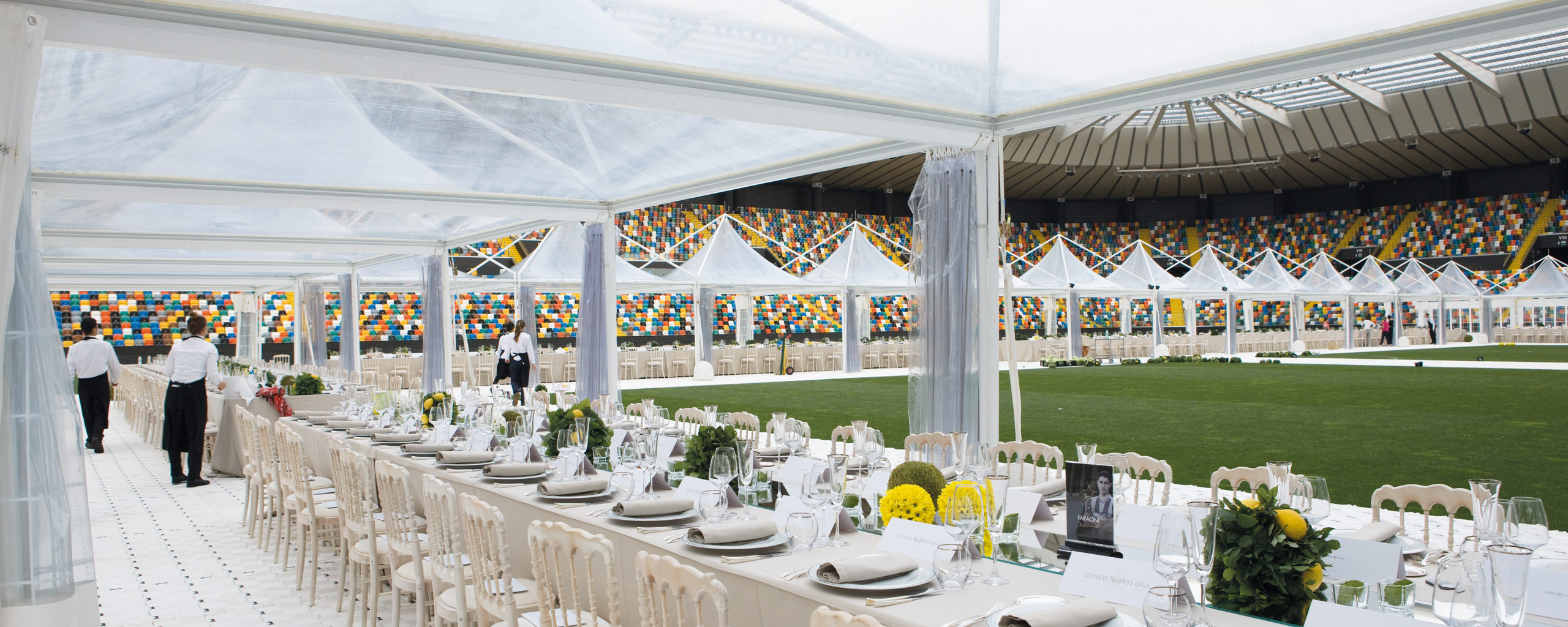 Un elegante banchetto all'aperto in uno stadio, con lunghi tavoli con tovaglie bianche, piatti e composizioni floreali. Lo spazio è coperto da una tenda trasparente e sullo sfondo sono visibili i colorati posti a sedere dello stadio.