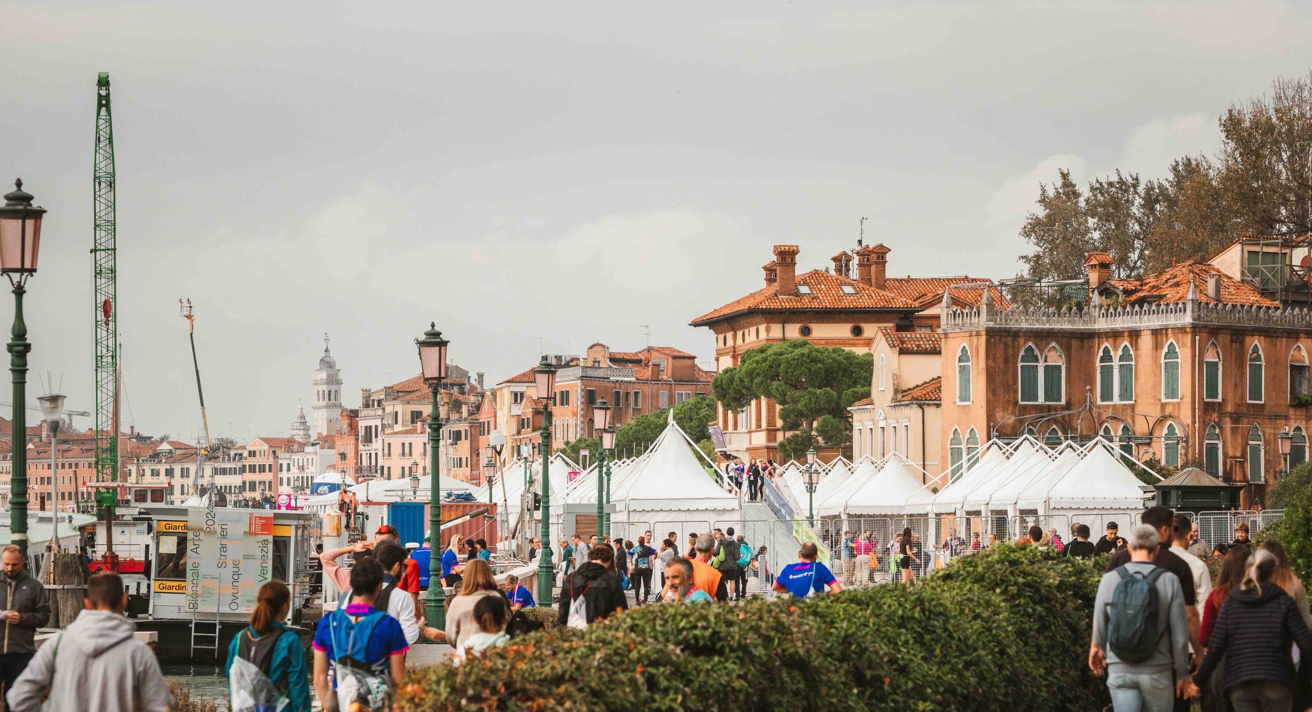 Una scena vivace a Venezia mostra una folla di persone che camminano lungo una passeggiata sul lungomare. Le tende costeggiano il sentiero e sullo sfondo sono visibili edifici storici con tetti rossi. Una gru si erge alta in mezzo alla pittoresca cornice.