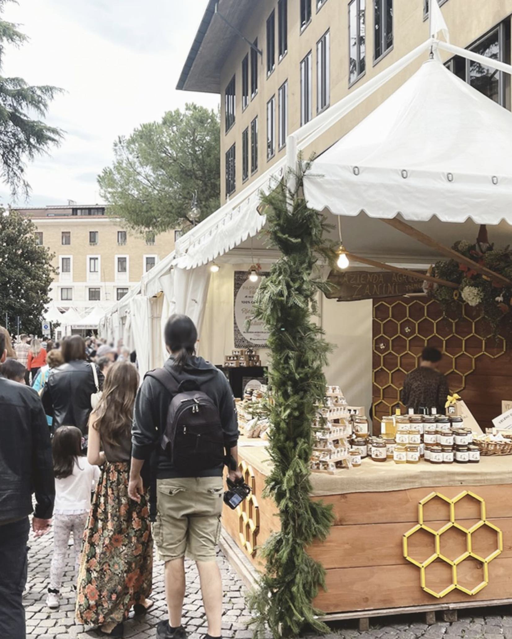 La gente curiosa in un vivace mercato all'aperto. Una bancarella adornata con un favo e vegetazione espone barattoli di miele. Le tende costeggiano il sentiero con la gente che si gode l'atmosfera vivace.