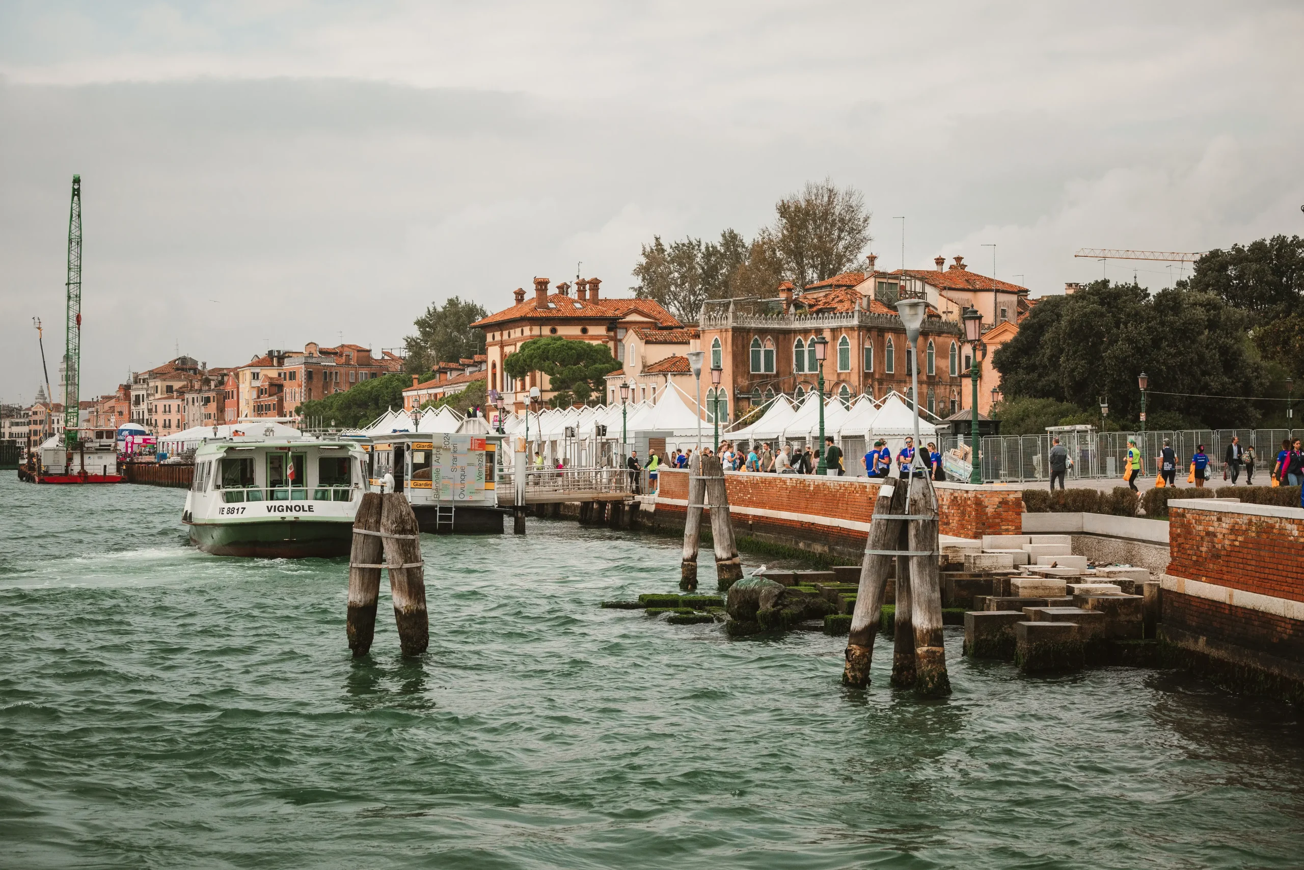 Una vista panoramica di un'area sul lungomare con un traghetto chiamato "Venezia" attraccato vicino a un molo. Ci sono edifici in mattoni, tende bianche e una fila di alberi sullo sfondo. Il cielo è coperto e l'acqua è di un verde agitato.