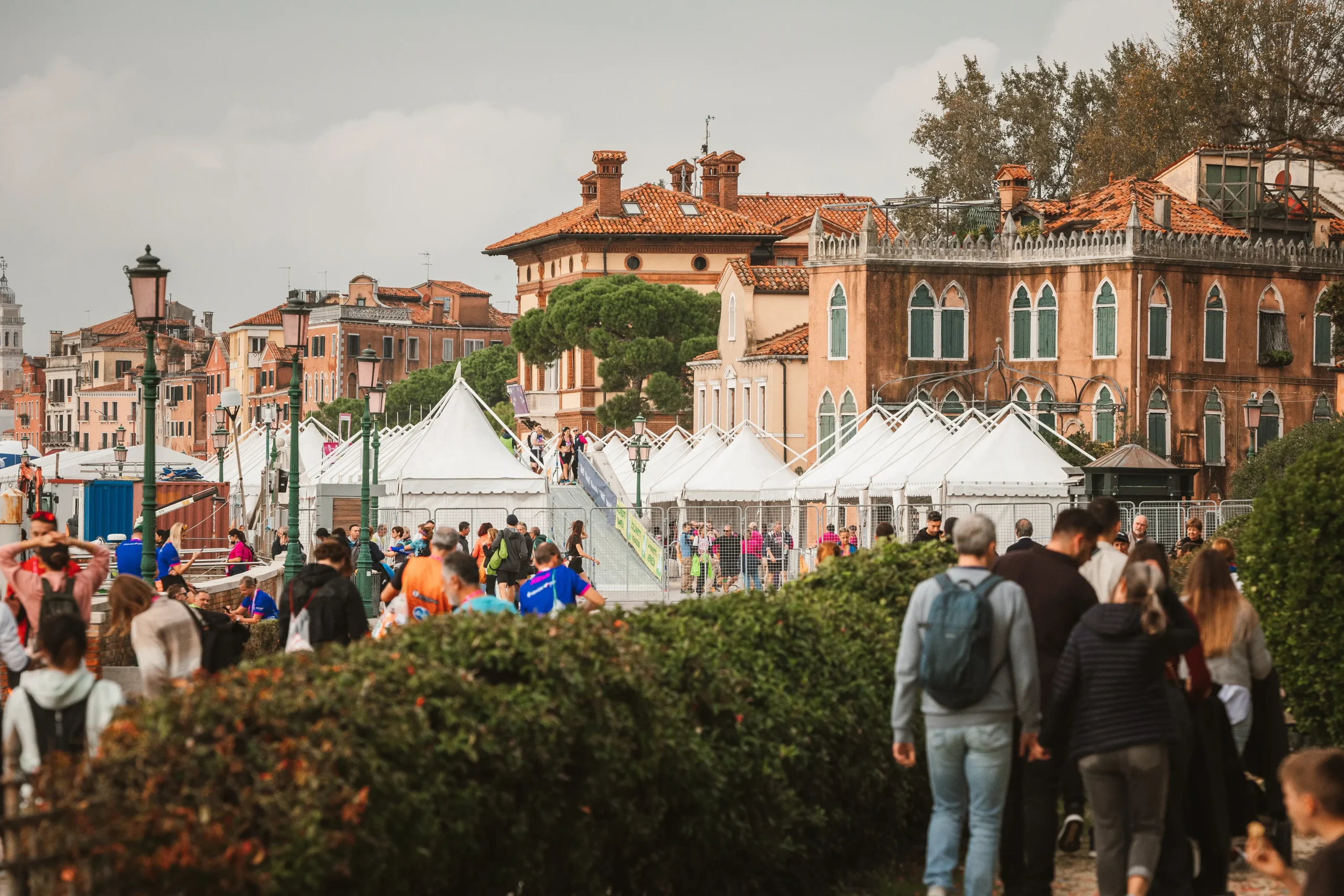 Persone che camminano lungo un sentiero costeggiato di verde in un'area urbana con edifici storici. Sullo sfondo sono montate tende bianche e in alto c'è un cielo nuvoloso.