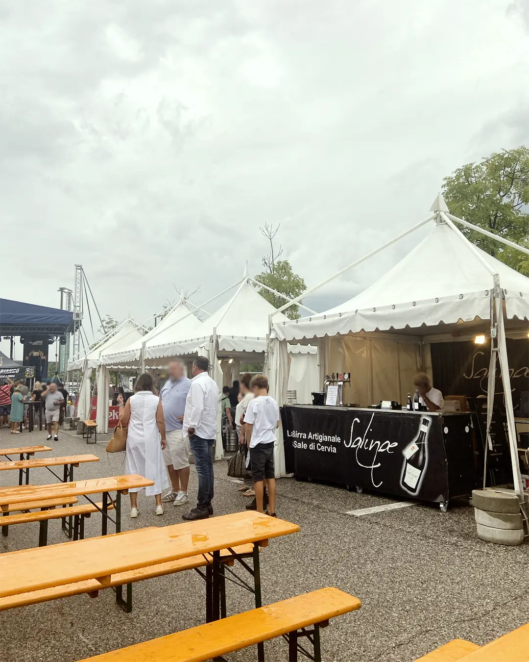 Festival gastronomico all'aperto con tende bianche e panche di legno. Le persone sono radunate attorno a una bancarella con l'etichetta "Salina", sotto cieli nuvolosi. Gli alberi sono visibili sullo sfondo.