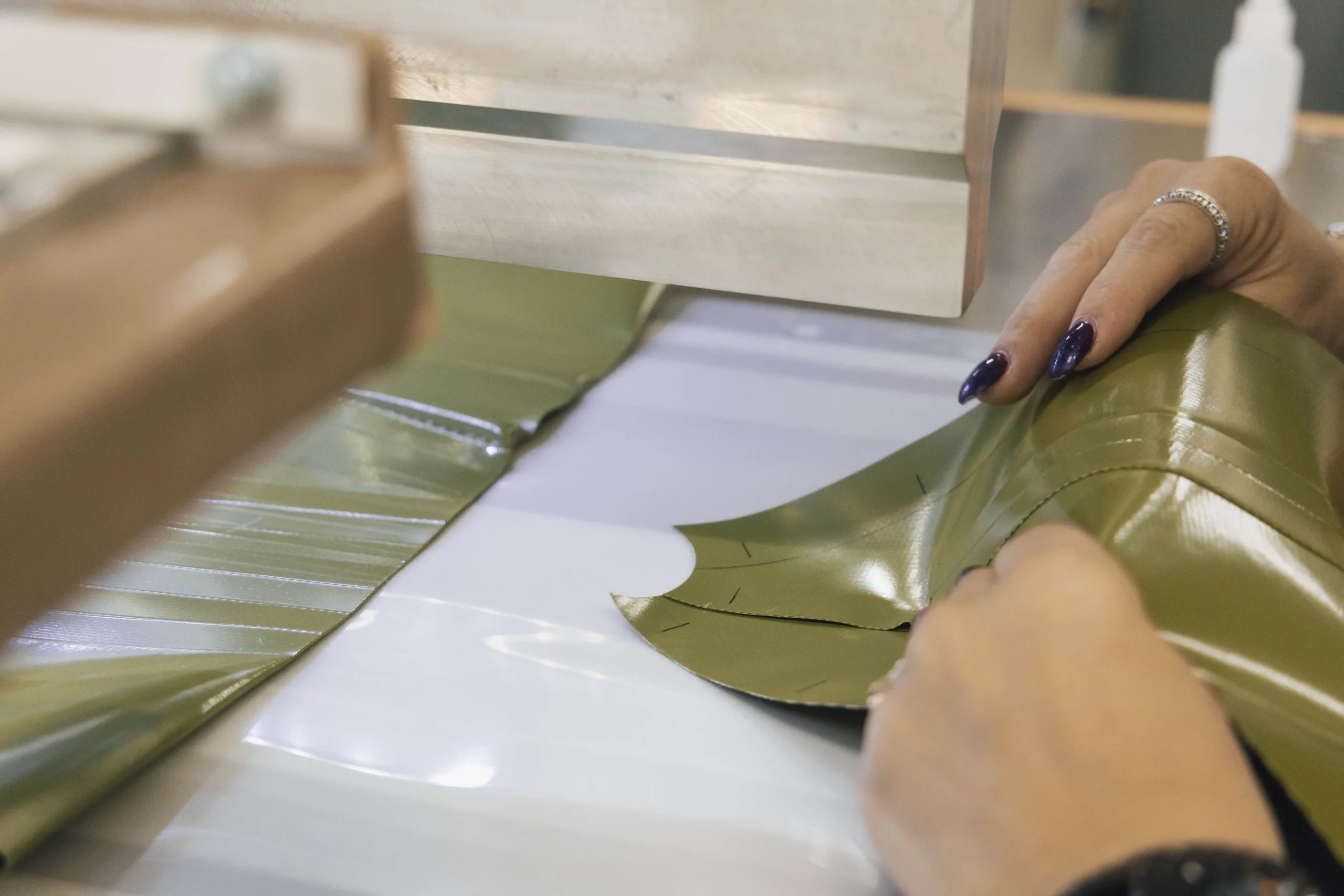 Una persona con smalto scuro e un anello in una mano lavora con un tessuto vinilico verde sotto una macchina da cucire. La persona guida il tessuto, che ha delle marcature, mentre passa attraverso la macchina.