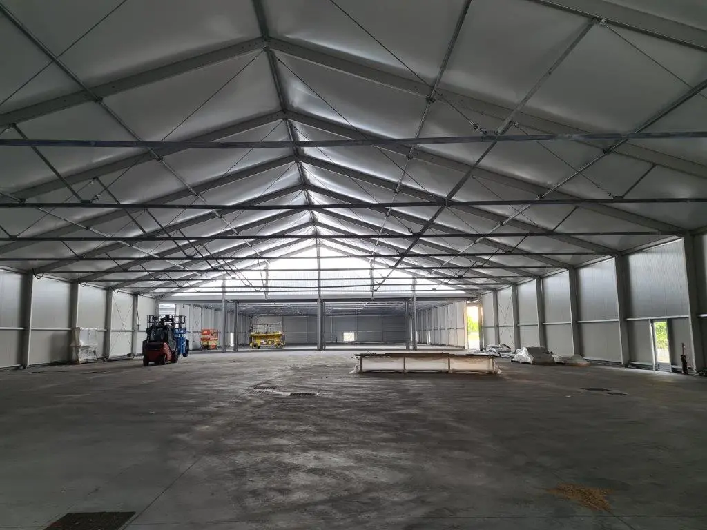 Vista interna di un grande magazzino vuoto con un soffitto alto e triangolare sostenuto da travi metalliche. Un carrello elevatore è parcheggiato sulla sinistra e alcune attrezzature e materiali sono sparsi sul pavimento, con la luce che entra dalle finestre.