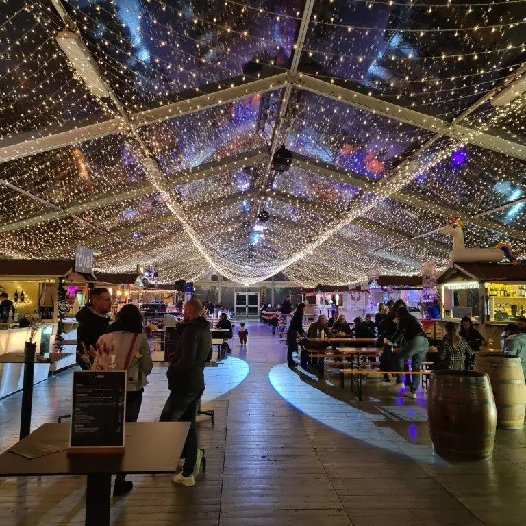 Un vivace mercato alimentare al coperto con un soffitto trasparente decorato con luci a corda, che creano un effetto stellato. Le persone siedono a tavoli di legno e si godono i pasti. Una varietà di bancarelle di cibo si allineano ai lati e un barile è in primo piano.