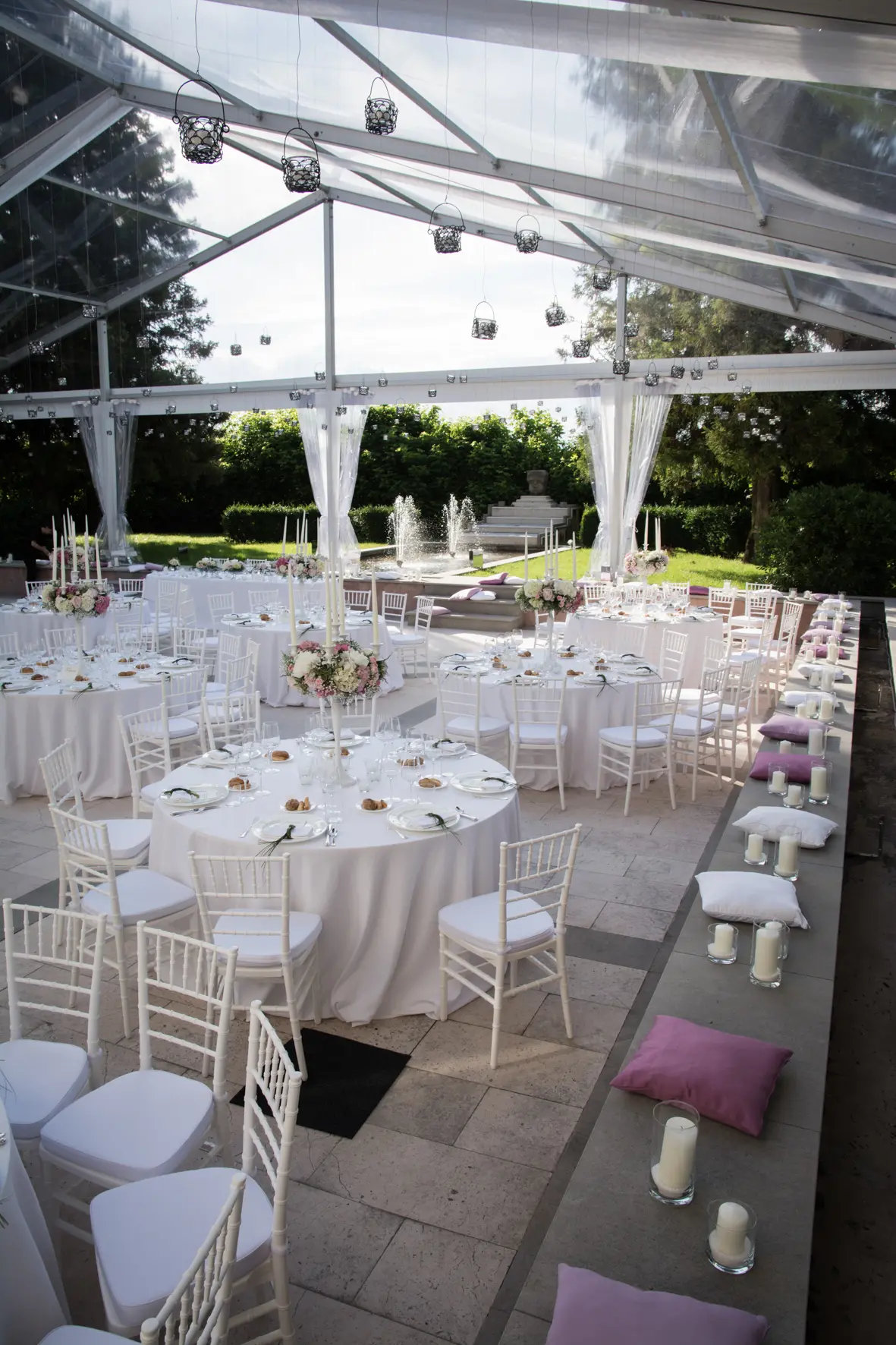 Elegante ricevimento di nozze all'aperto sotto una tenda trasparente, con tavoli rotondi con tovaglie bianche, centrotavola floreali e sedie bianche. Cuscini rosa costeggiano una sporgenza di pietra, con candele nelle vicinanze. Una fontana e una vegetazione lussureggiante sono visibili sullo sfondo.