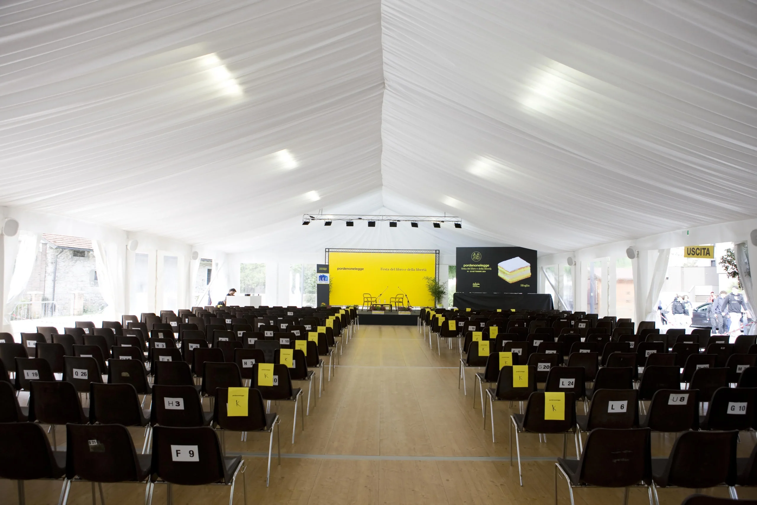 Una spaziosa tenda per eventi con file di sedie marroni di fronte a un palco. Il palco ha due sedie vuote ed è decorato con uno sfondo giallo e nero, che mostra un disegno di un libro. Luci luminose e uniformemente distanziate illuminano l'interno bianco.