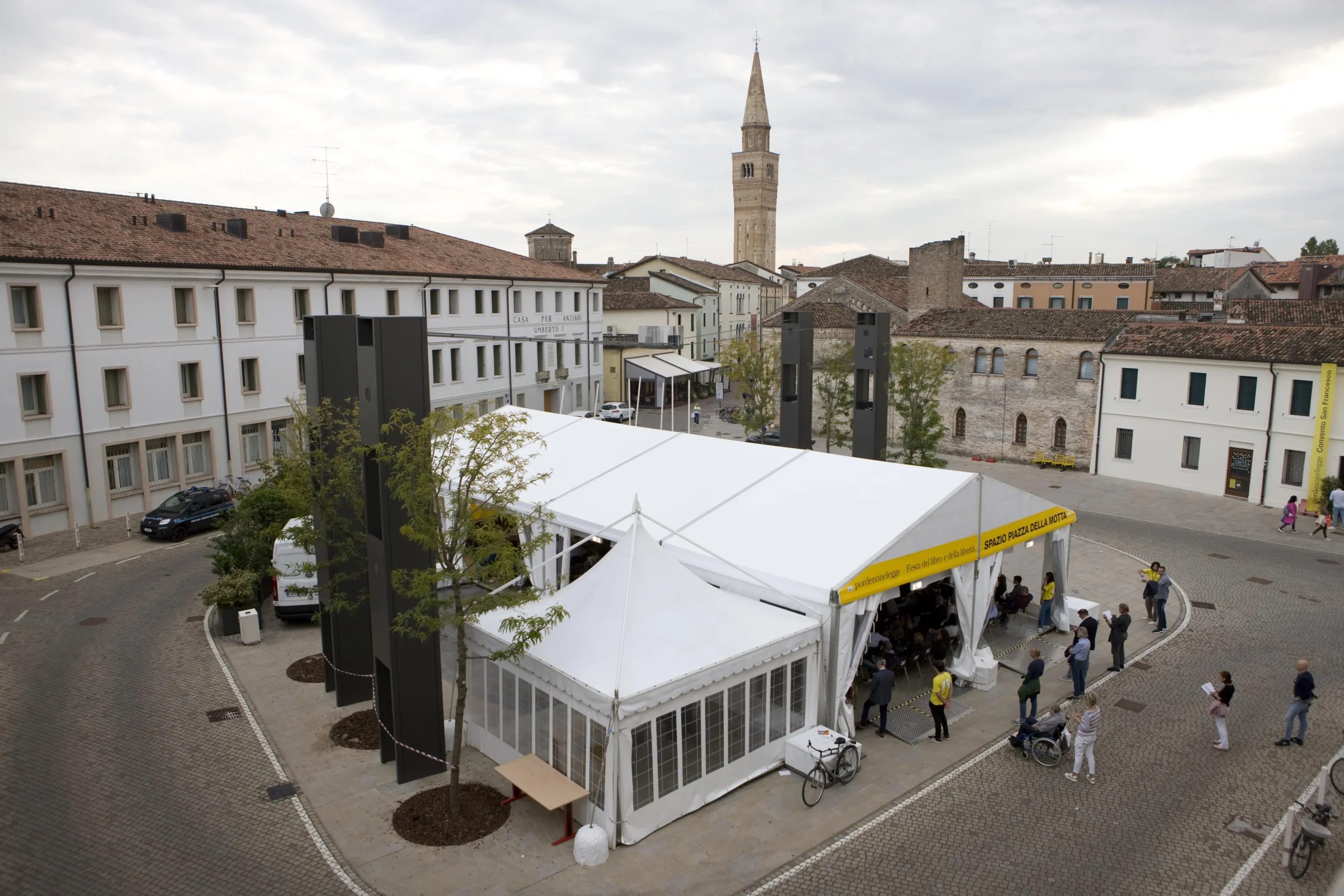 Vista aerea di una tenda bianca allestita in una piazza acciottolata circondata da edifici storici. La gente si raduna vicino all'ingresso. Un alto campanile è visibile sullo sfondo sotto un cielo nuvoloso.