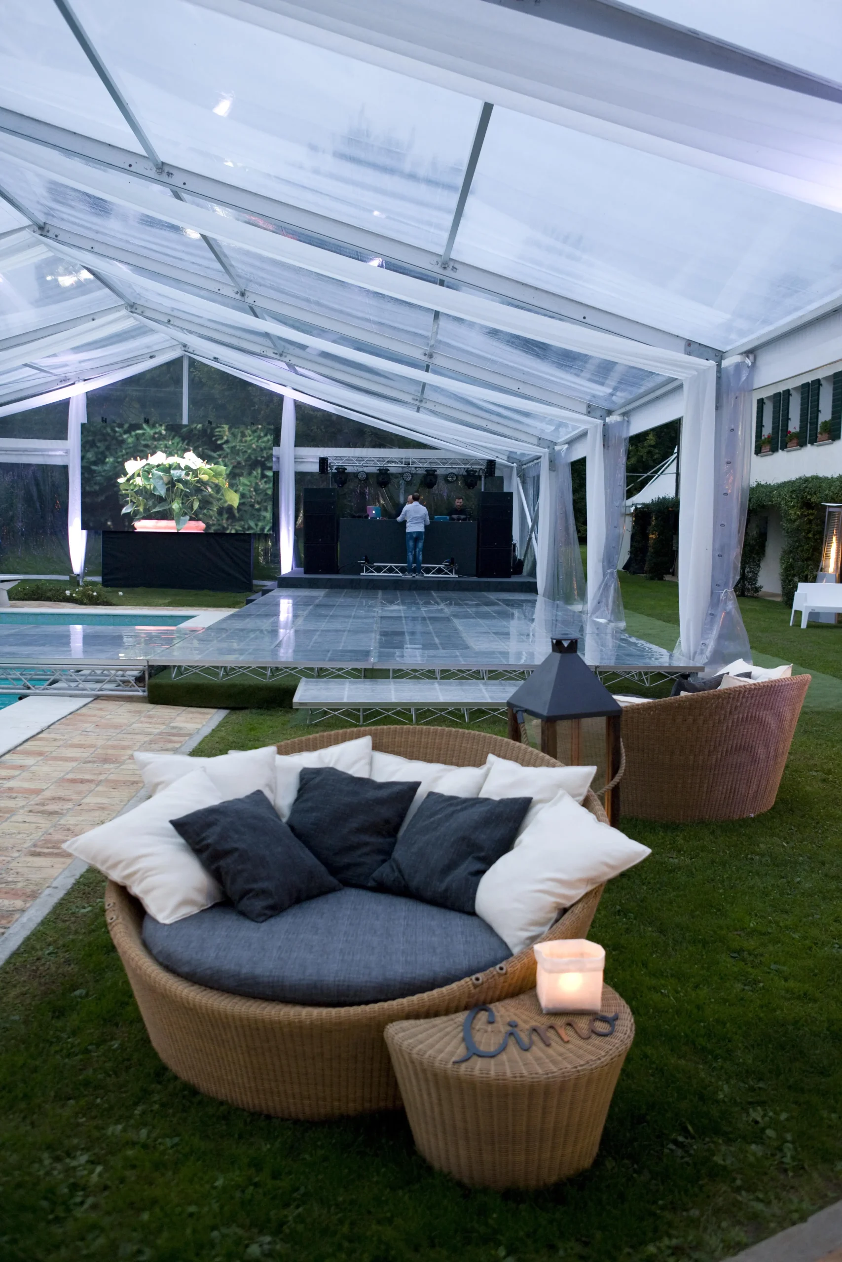 Un'elegante configurazione esterna con una tenda trasparente con pista da ballo e postazione DJ. Un comodo divano circolare in vimini con cuscini grigi è in primo piano e l'area è circondata da erba e alberi, creando un'atmosfera accogliente per un evento.