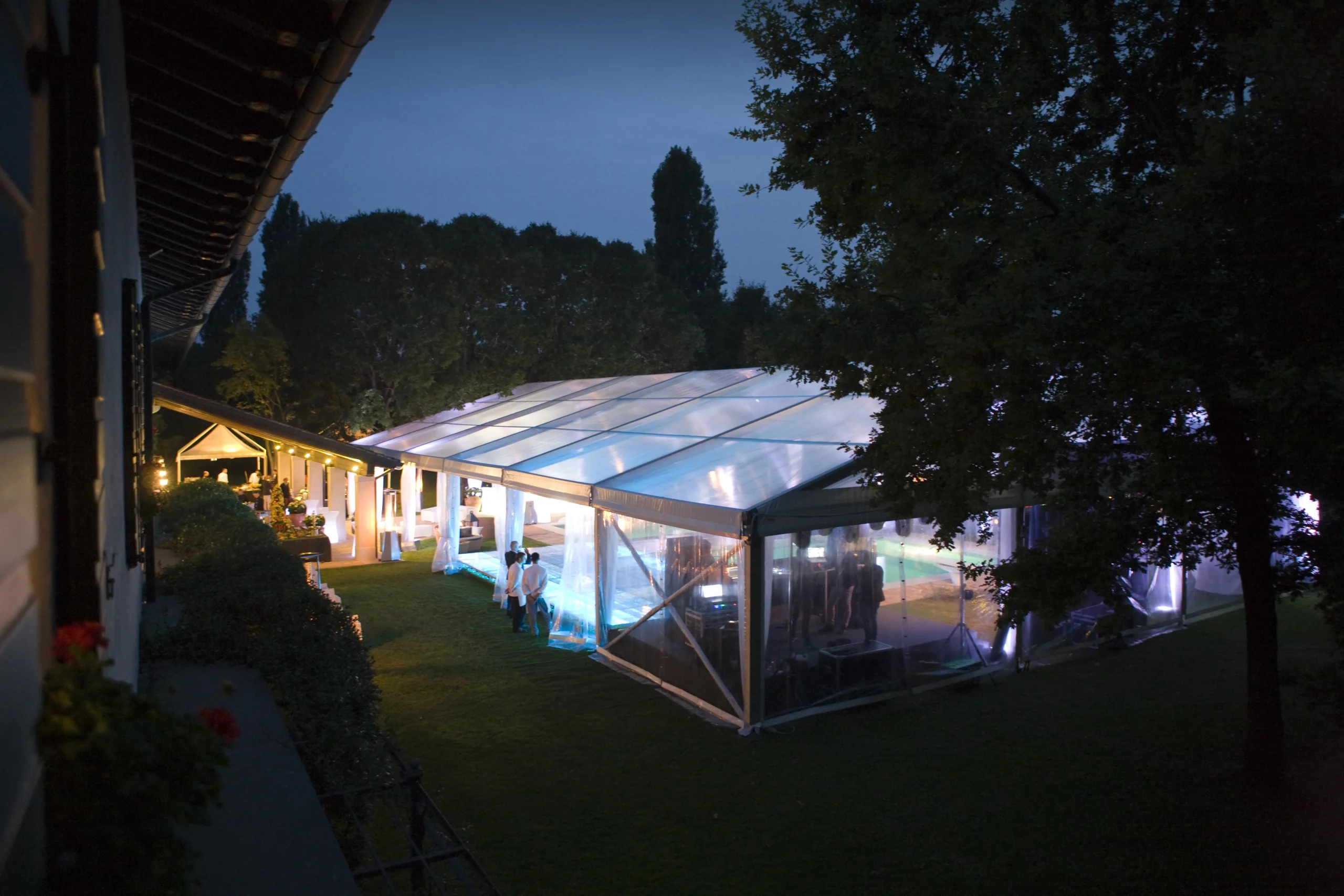 Una scena notturna di una grande tenda trasparente allestita all'aperto, che brilla di luci soffuse viola e blu. Gli alberi circondano l'area e un edificio vicino è scarsamente illuminato. Sono visibili delle persone all'interno della tenda, il che suggerisce un evento o un raduno.