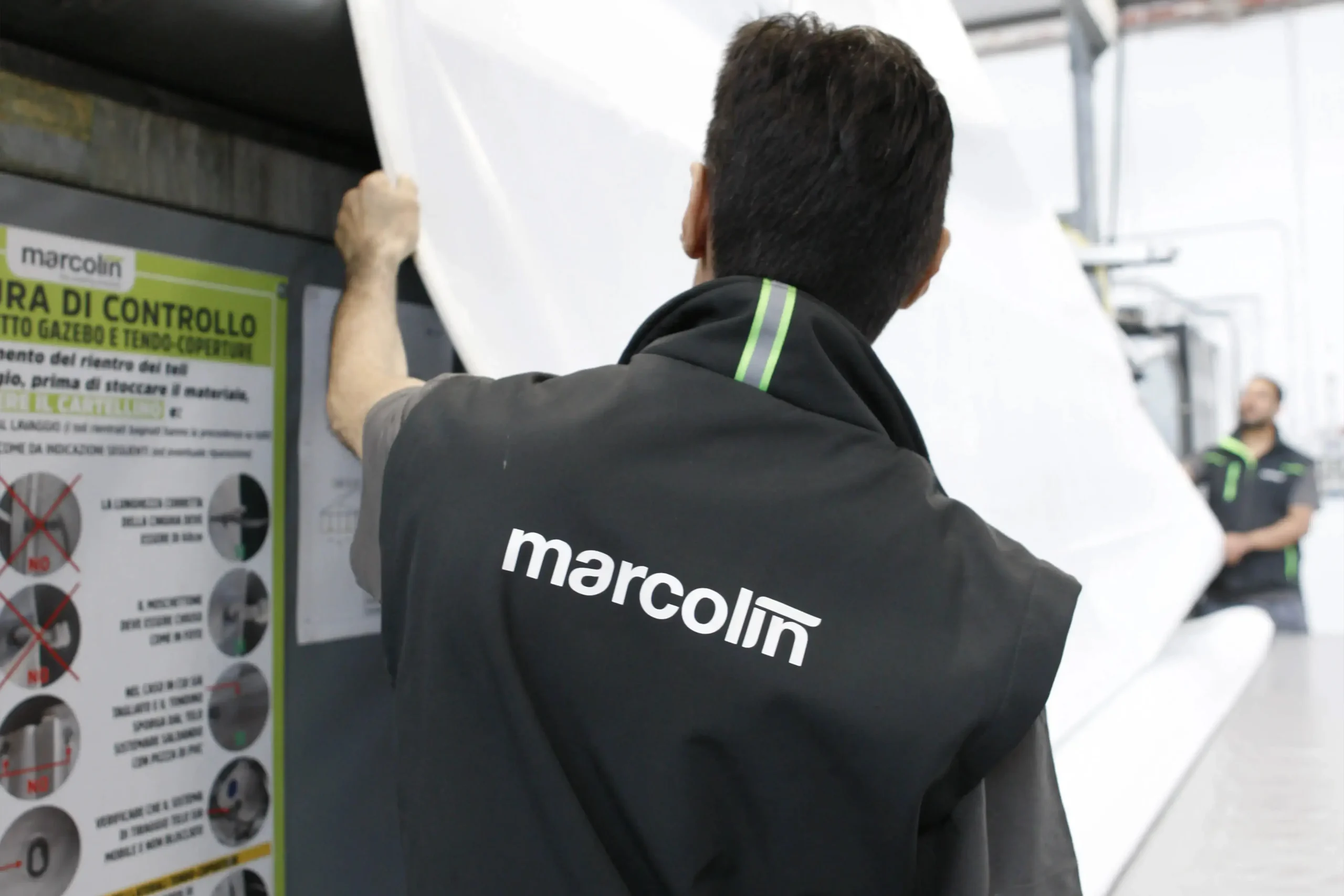 Una persona che indossa un gilet con la scritta "marcolin" sulla schiena sta sistemando un grande telo di tessuto bianco in un ambiente industriale. Un'altra persona è visibile sullo sfondo, anche lei con un abbigliamento simile.