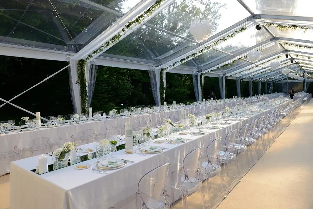 Un lungo ed elegante ricevimento di nozze allestito sotto una tenda trasparente. I tavoli sono coperti da tovaglie bianche, decorati con fiori bianchi e candele. Sedie trasparenti allineate lungo i tavoli e l'ambiente è circondato da una vegetazione lussureggiante.