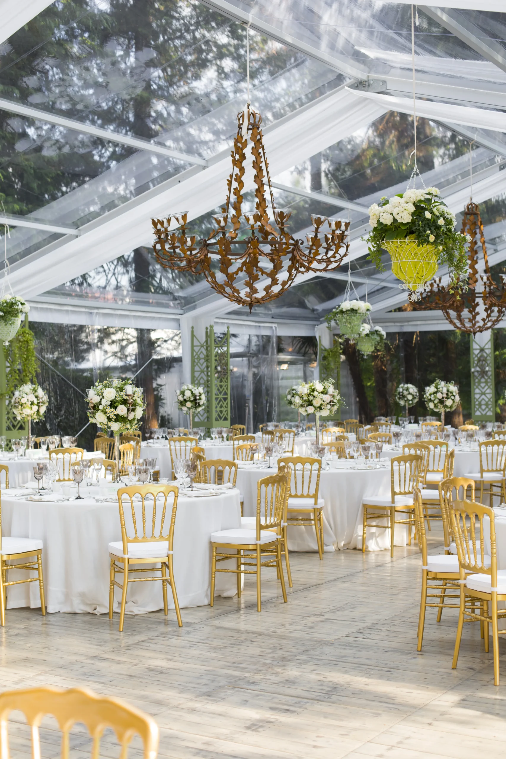 Elegante ricevimento di nozze con tavoli drappeggiati di bianco, sedie dorate e sontuosi centrotavola floreali sotto una tenda trasparente. Grandi lampadari decorati pendono dal soffitto, illuminando l'atmosfera sofisticata e ariosa.