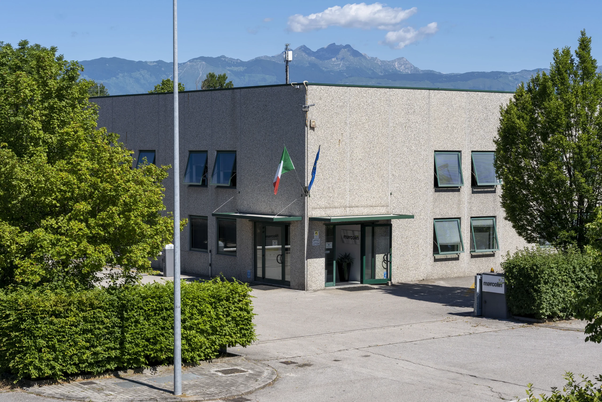 Un edificio grigio a due piani con bandiere italiane e dell'Unione Europea accanto all'ingresso. I cespugli circondano l'area e le montagne sono visibili sullo sfondo sotto un cielo azzurro terso.
