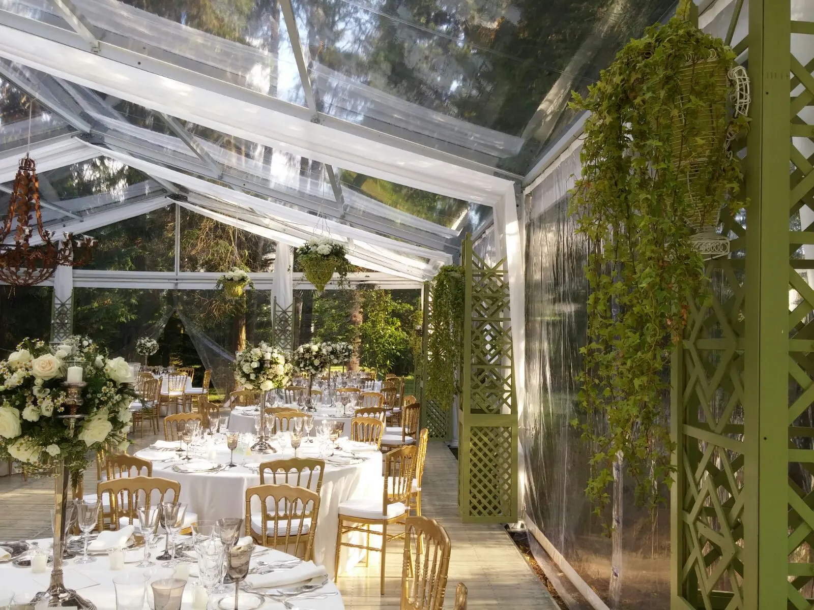 Un ricevimento di nozze sotto una tenda trasparente, decorato con verde pensile e composizioni floreali. I tavoli rotondi sono apparecchiati con tovaglie bianche, eleganti stoviglie e sedie. La calda luce del sole entra a fiotti, creando un'atmosfera serena e festosa.