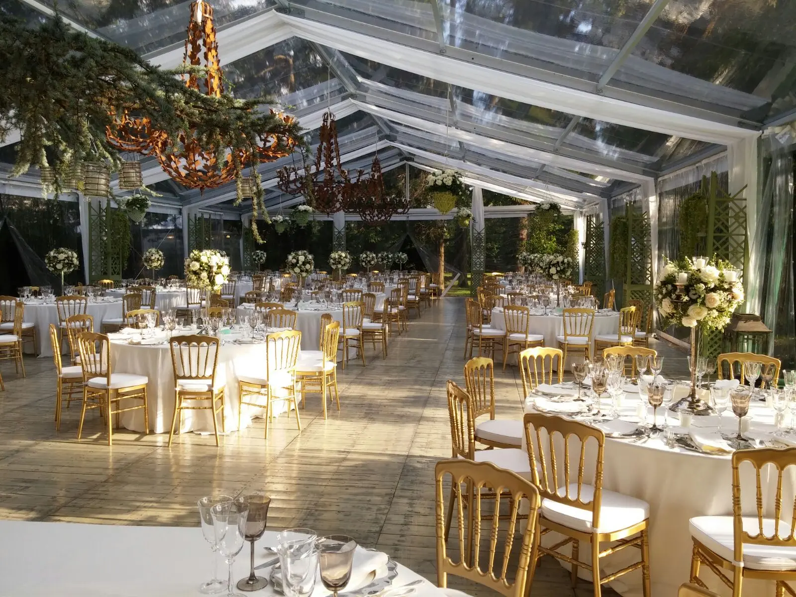 Elegante location per matrimoni allestita in una tenda trasparente con tavoli rotondi decorati con tovaglie bianche, sedie dorate e centrotavola floreali. Lampadari pendono dal soffitto e la luce del sole filtra, creando un'atmosfera calda e invitante.