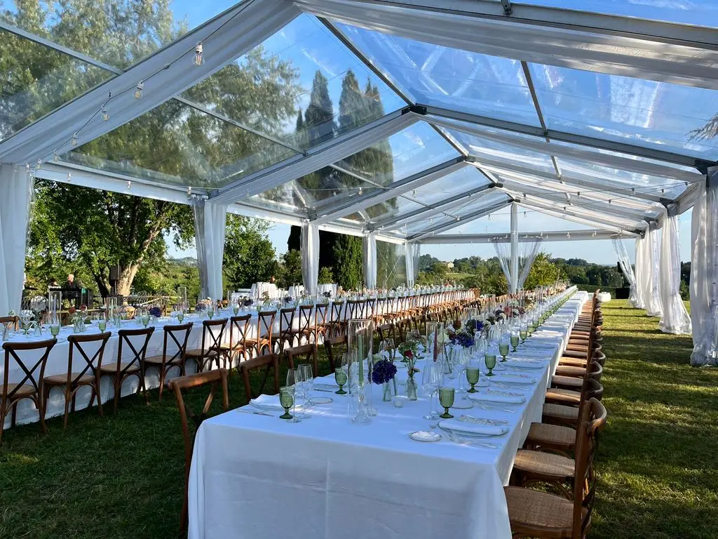 Lunghi tavoli con tovaglie bianche sono sistemati sotto una tenda trasparente all'aperto, circondati dal verde. Le sedie fiancheggiano i tavoli, che sono adornati con bicchieri e centrotavola floreali, creando un'elegante cornice per l'evento.