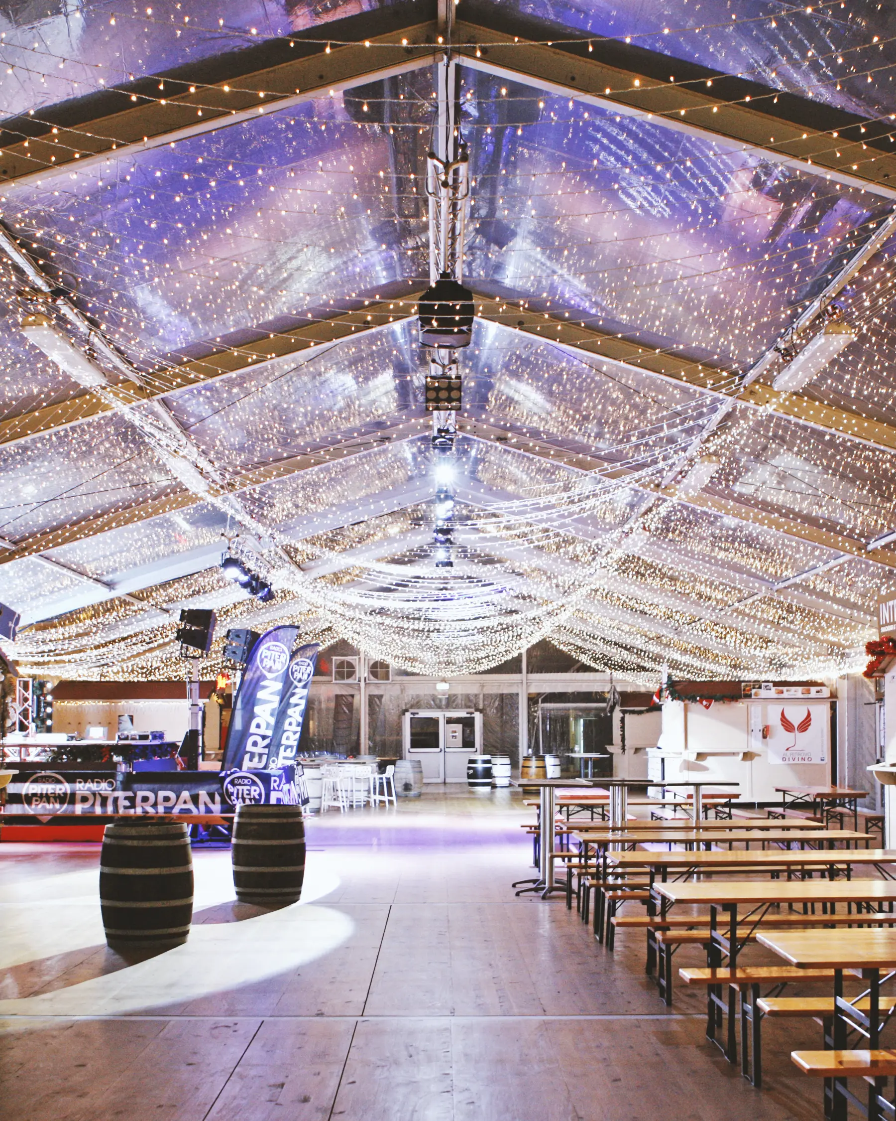 Una spaziosa tenda per eventi con tetto e soffitto trasparenti, adornati da lucine scintillanti. All'interno ci sono tavoli da picnic in legno e decorazioni a forma di botte. L'area è ben illuminata, creando un'atmosfera festosa.