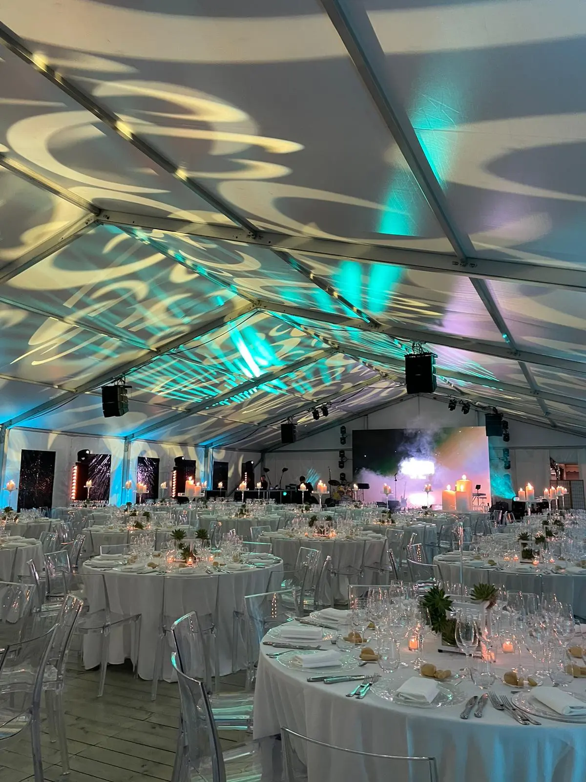 Un grande ed elegante tendone per eventi con tavoli rotondi coperti da tovaglie bianche, apparecchiati con bicchieri e posate. In alto, l'illuminazione artistica crea motivi sul soffitto interno e un palco con strumenti è visibile all'estremità.