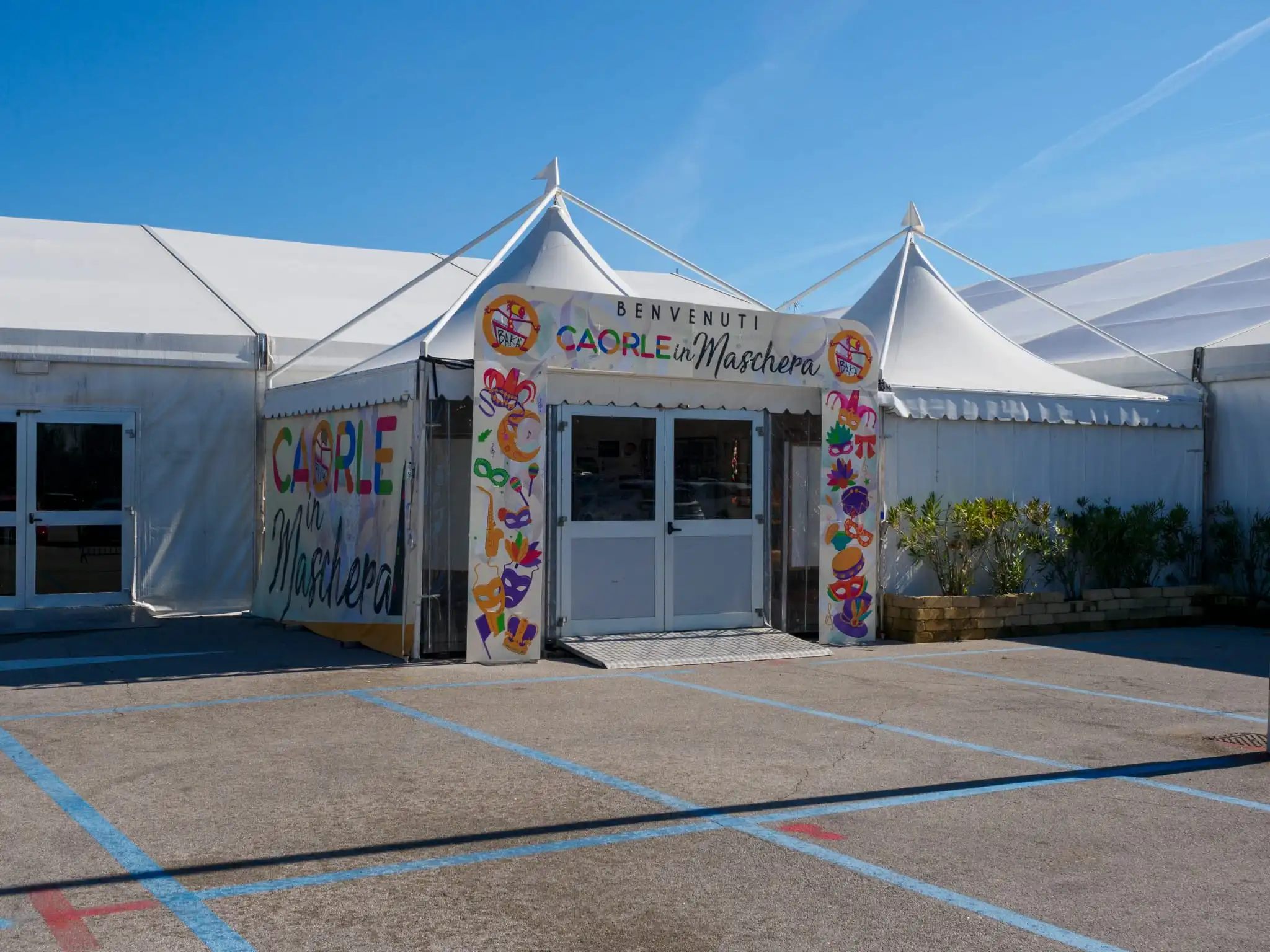 Una tenda bianca per eventi con decorazioni colorate e cartelli con la scritta "Benvenuti" e "Caorle nelle Marche". La tenda si trova in un parcheggio con un cielo azzurro e limpido sopra. Ci sono piccole piante vicino all'ingresso.