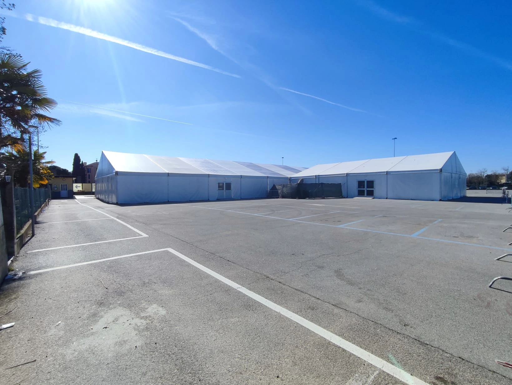 Un'ampia area esterna con grandi tende bianche montate in una giornata di sole. Il terreno è pavimentato, con linee visibili, che suggeriscono parcheggi o sentieri. Un cielo azzurro e terso è in alto, e si possono vedere alcuni alberi sparsi intorno.