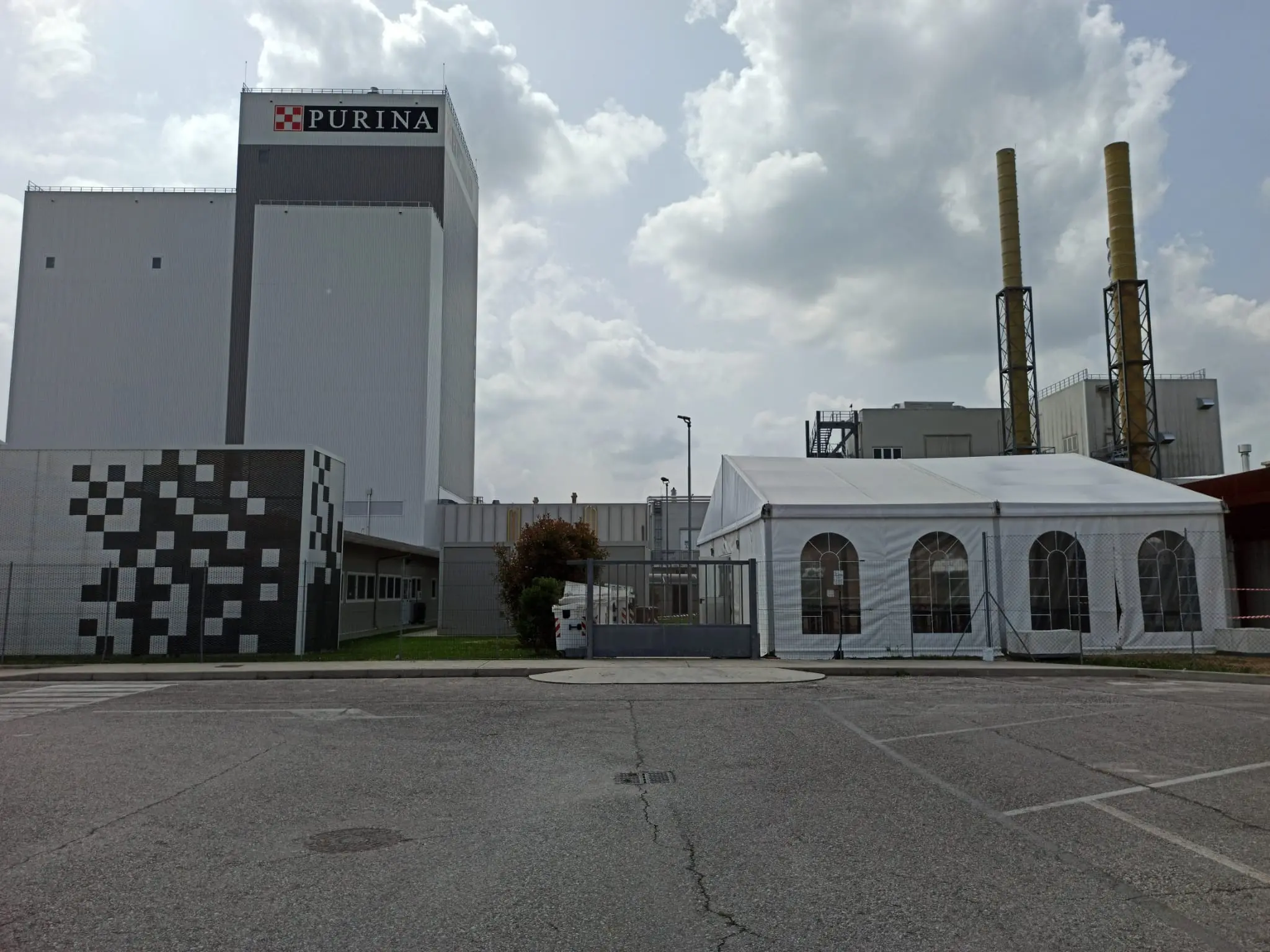 È visibile un grande edificio industriale con il logo Purina. Sulla destra, una tenda bianca è montata vicino a due alte ciminiere. Il cielo è per lo più nuvoloso. In primo piano c'è un parcheggio vuoto.